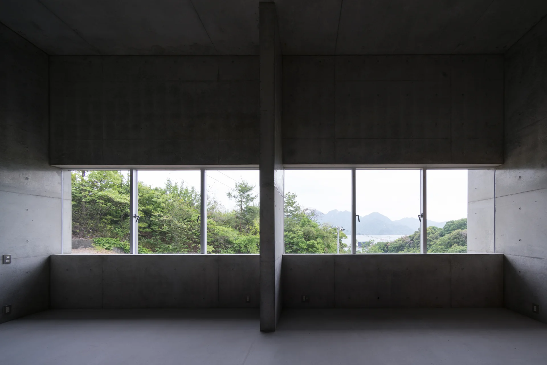 An empty concrete interior featuring raw grey concrete walls, a central structural pillar, and wide horizontal windows looking out at green hills.