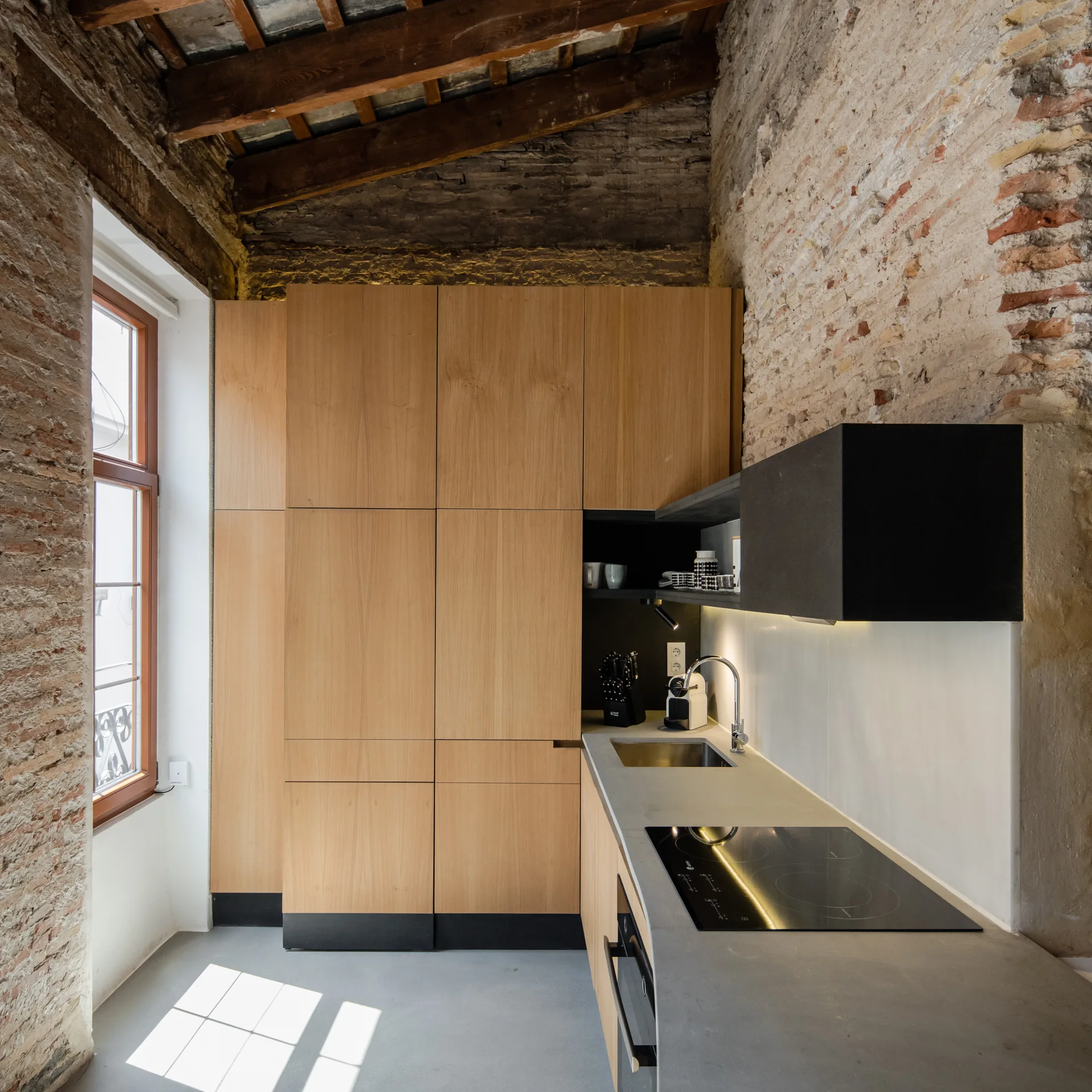 A modern kitchen featuring flat-front wood cabinets, concrete countertops, a built-in cooktop, exposed brick walls, and rustic timber ceiling beams.