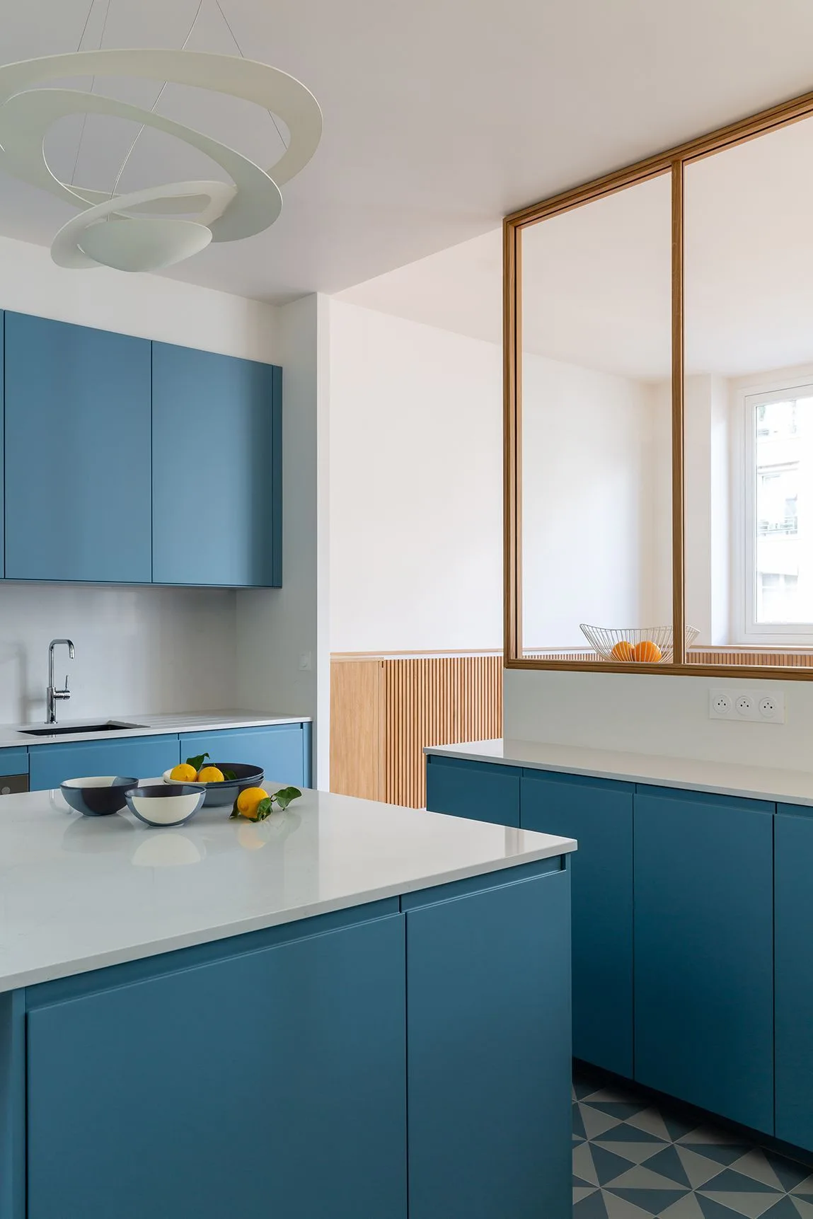 A modern kitchen featuring matte blue cabinetry, white countertops, geometric floor tiles, and a wood-framed glass partition wall.
