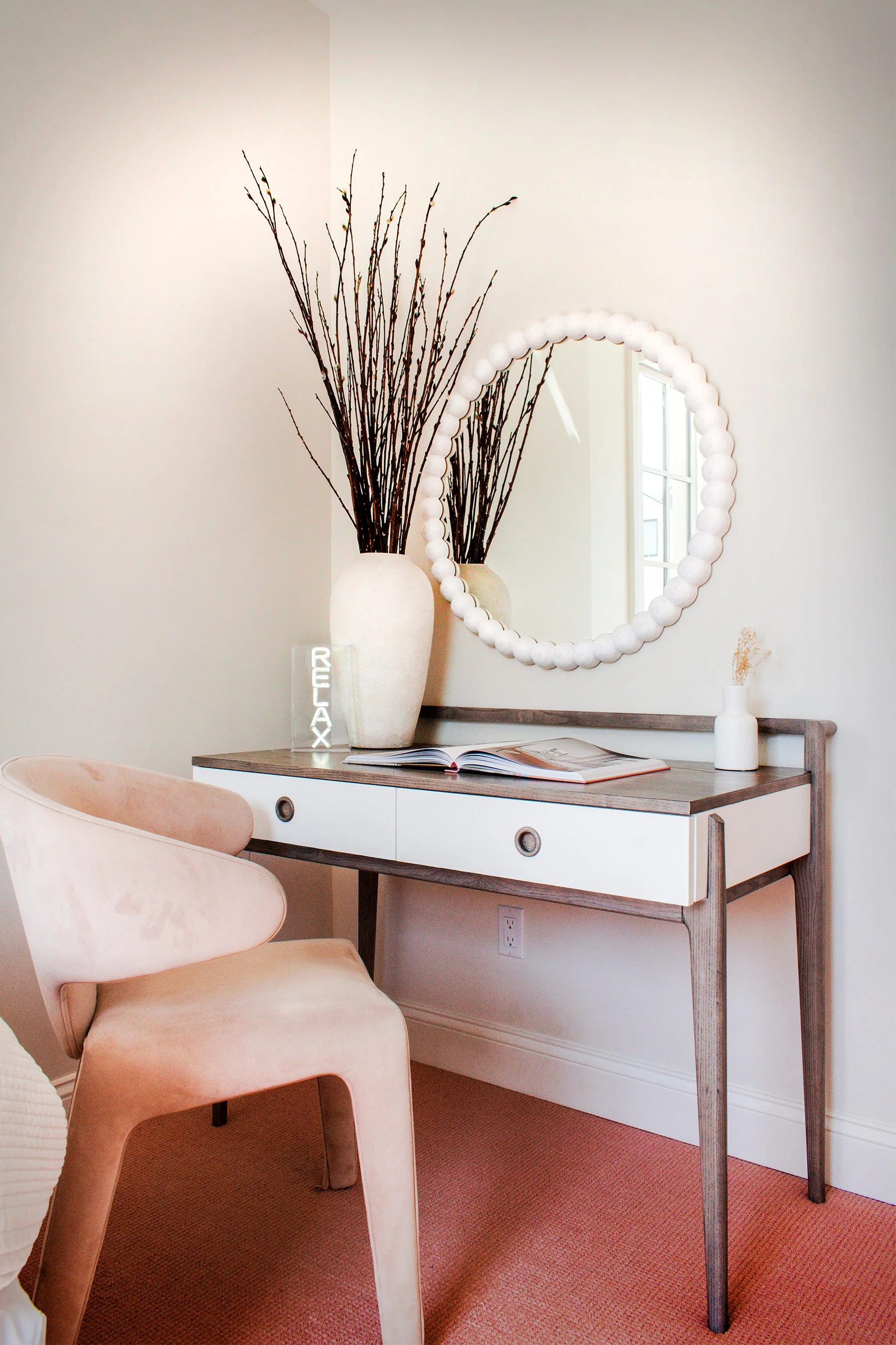 A modern two-tone writing desk features a round beaded mirror, a tall textured vase with branches, and an upholstered pink chair on carpet.