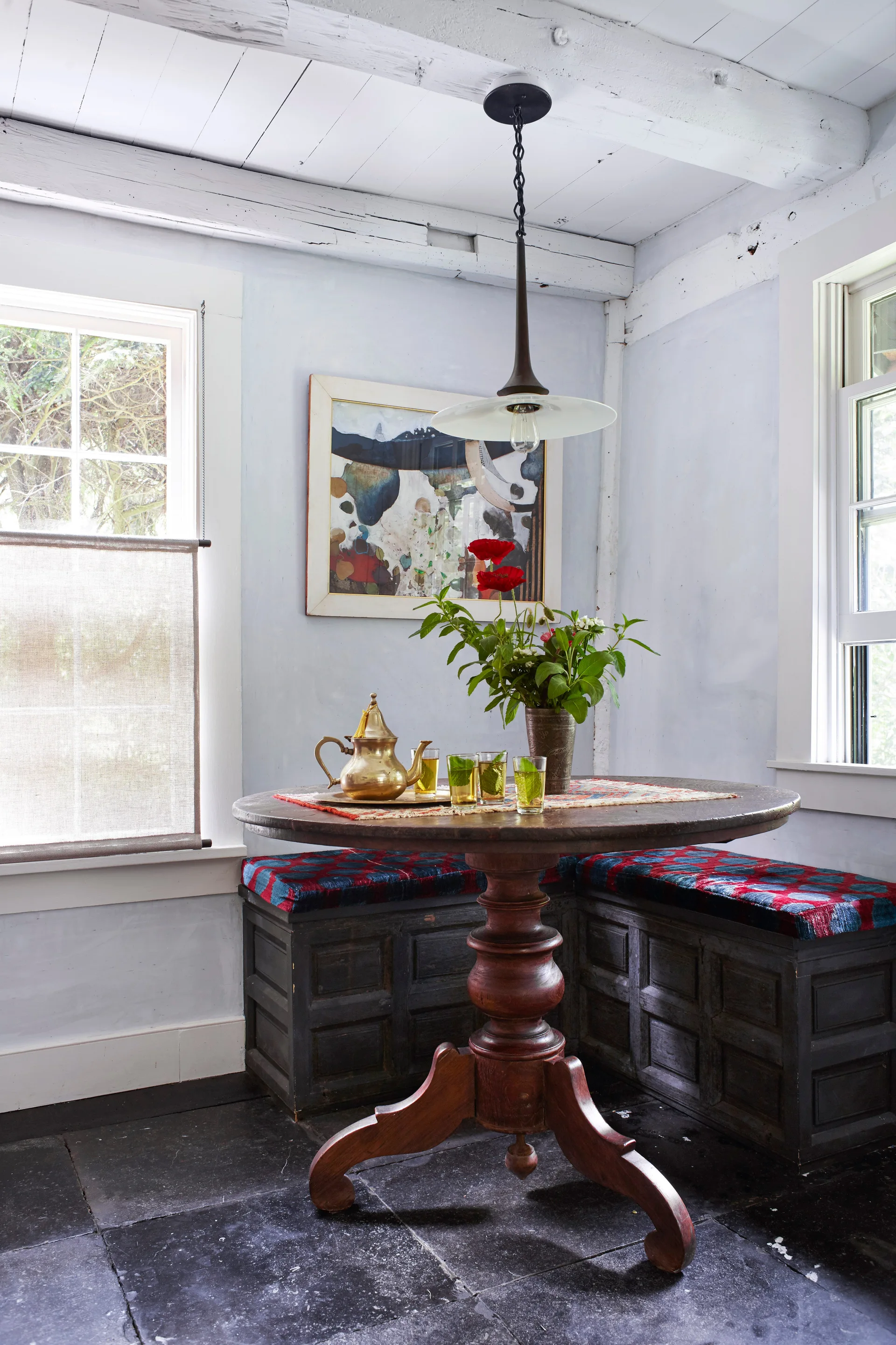 A rustic dining nook features a round wooden pedestal table, a built-in corner banquette with patterned cushions, and painted wood ceiling beams.