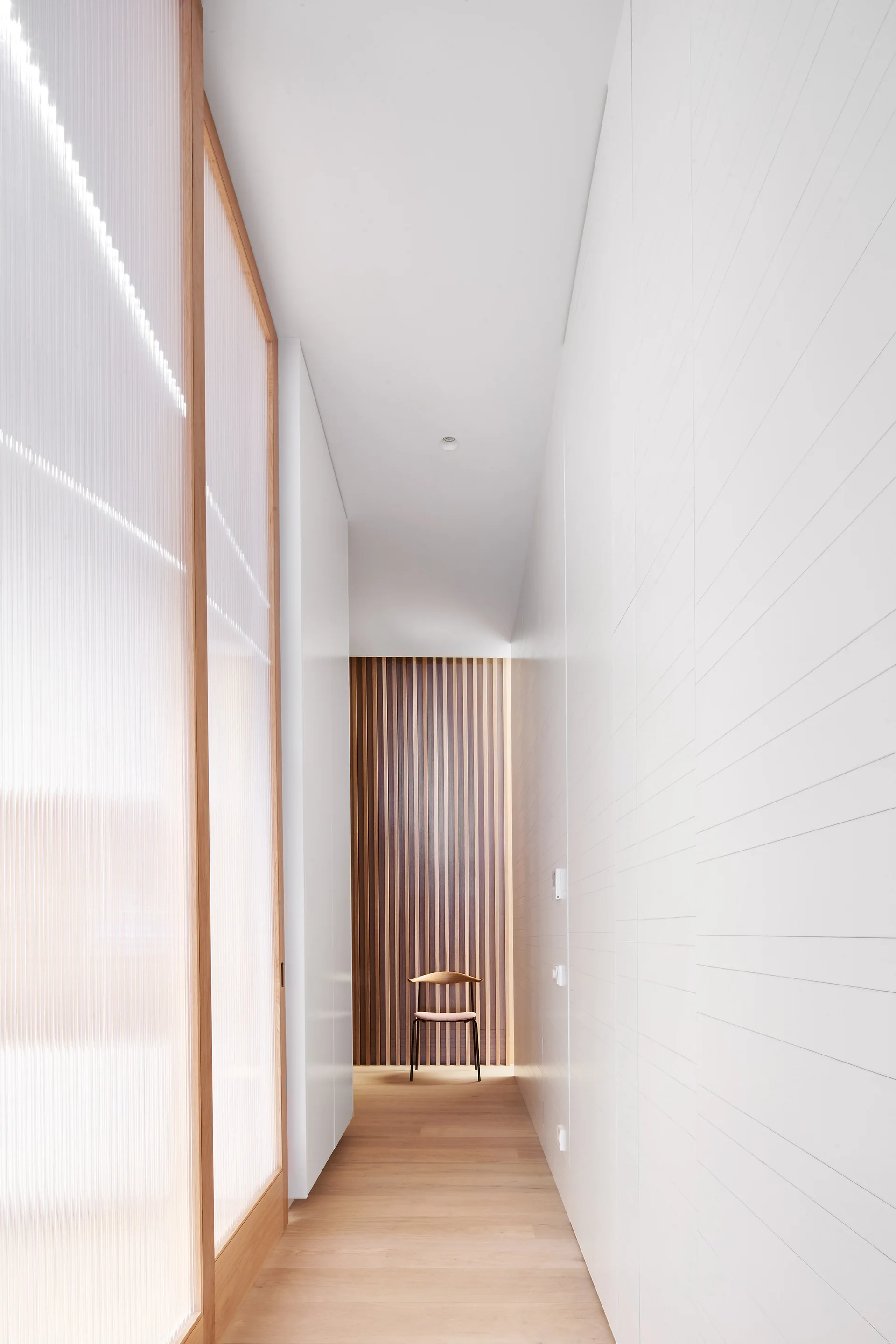 A minimalist hallway with white walls, light wood floors, a fluted glass partition, and a single wooden chair against a wood slat wall.
