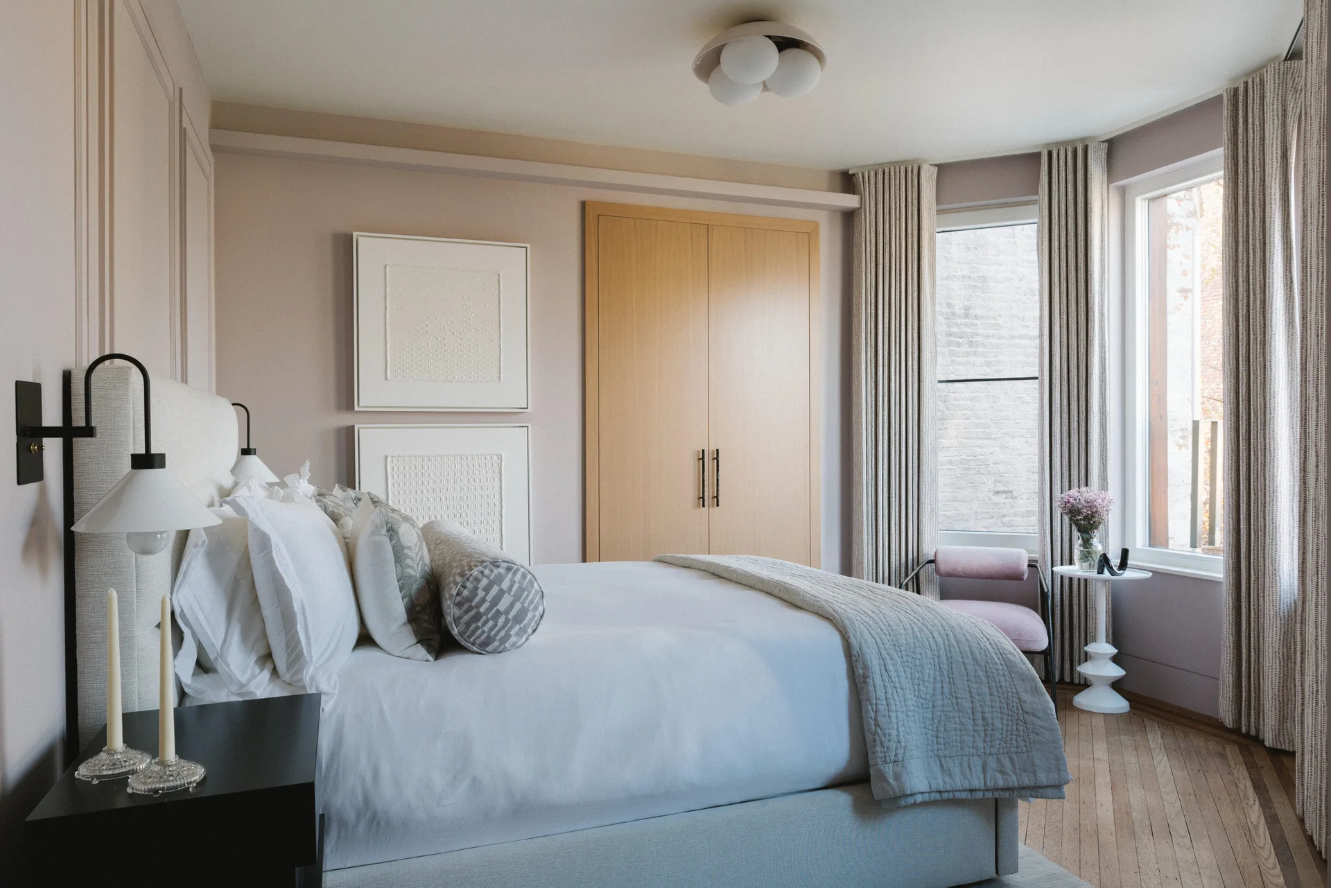 A modern bedroom features a bed with white bedding, mauve walls, a light wood closet, a pink chair, and striped curtains.