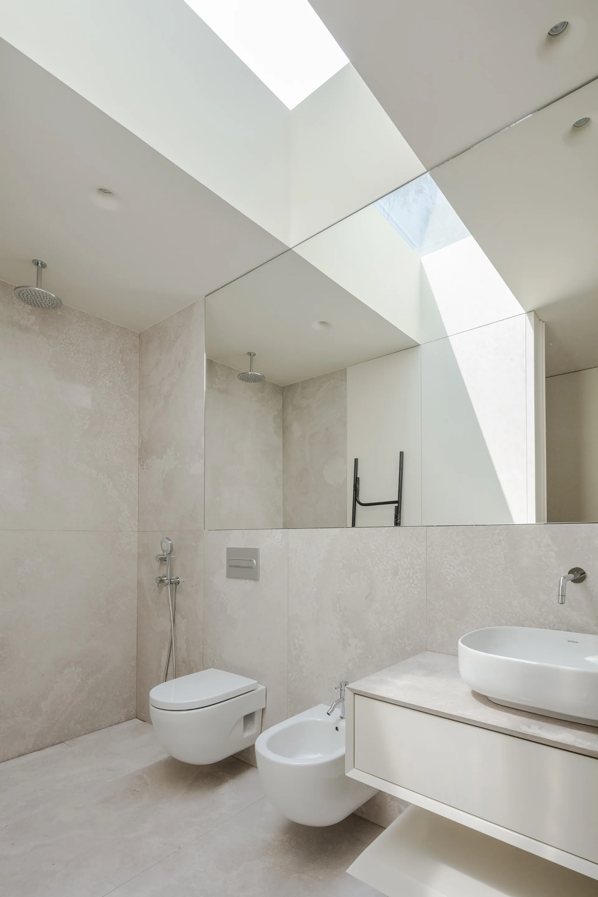 A minimalist beige bathroom features a large overhead skylight, a full-width wall mirror, a floating white vanity, and a wall-hung toilet.