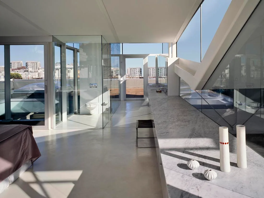 A modern bathroom featuring a long marble vanity, a glass-enclosed toilet stall, and large glass windows with a rooftop view.