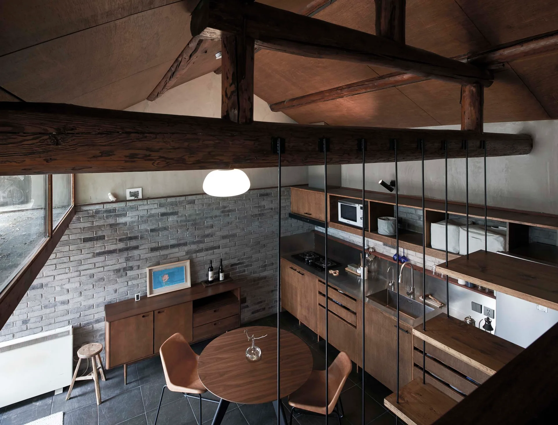 An overhead view of a rustic kitchen and dining area featuring a round wood table, exposed timber ceiling beams, and a grey brick wall.