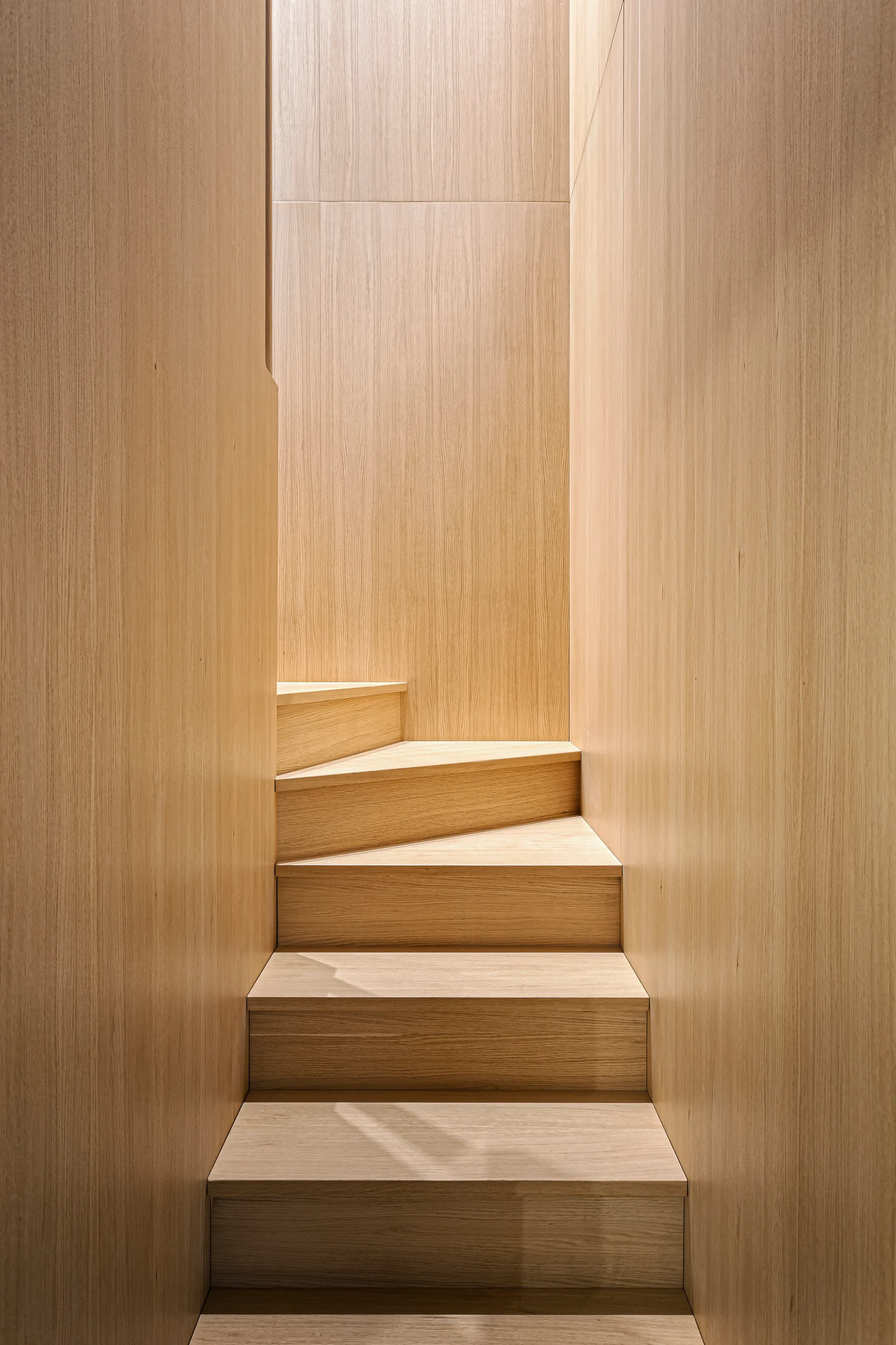 A minimalist wooden staircase winds upward between matching light wood paneled walls under soft overhead light.