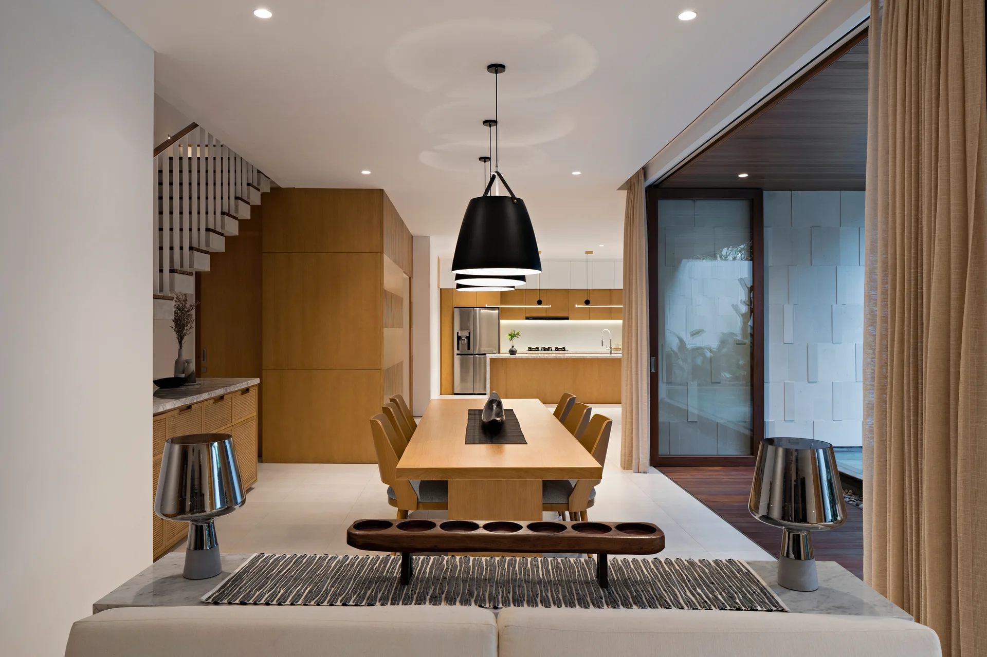 A contemporary open-plan dining room with a light wood table, oversized black pendant lights, and a kitchen visible in the background.