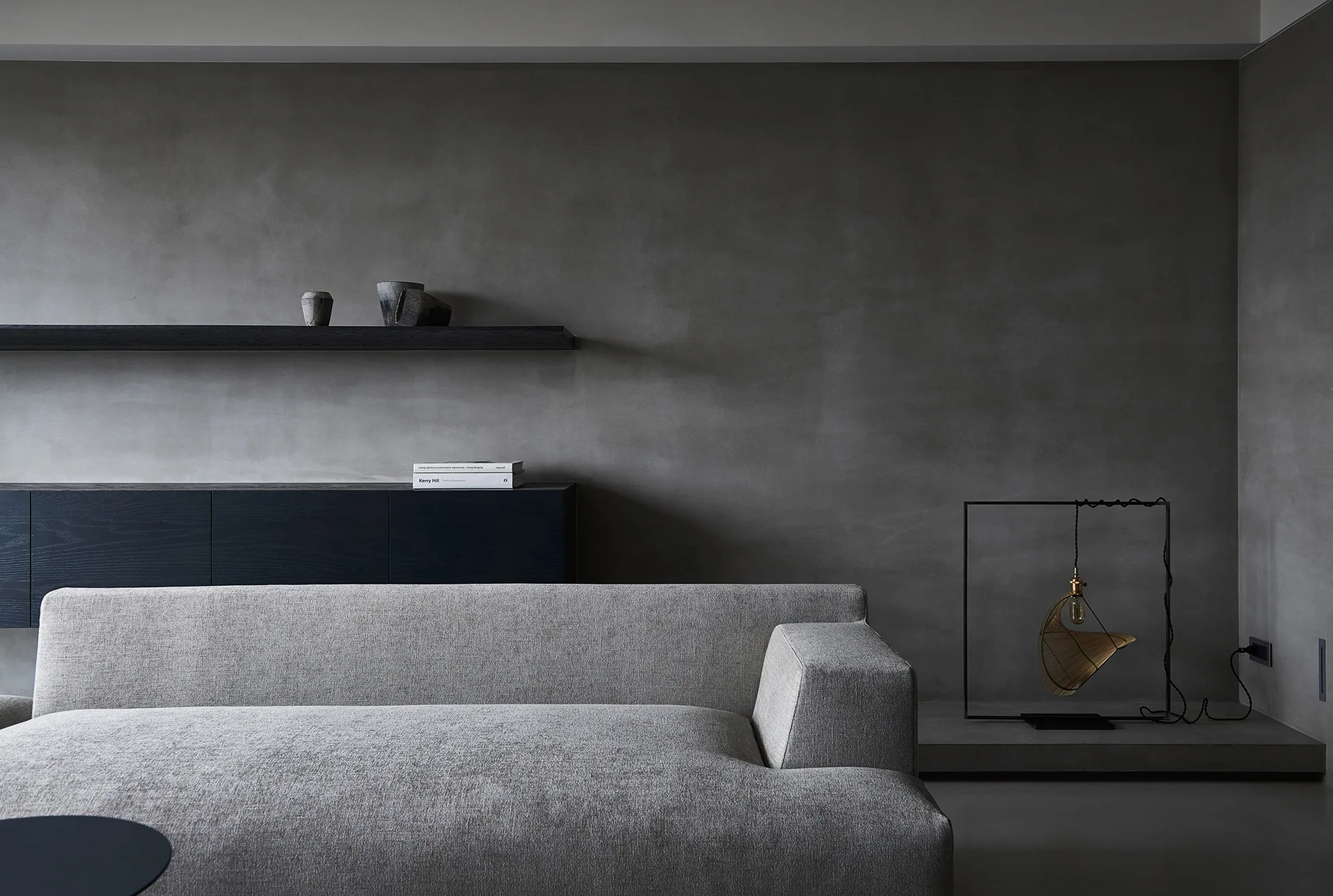 A light gray sofa sits against a dark gray plaster wall with a low black cabinet, floating shelf, and a geometric wire frame lantern.