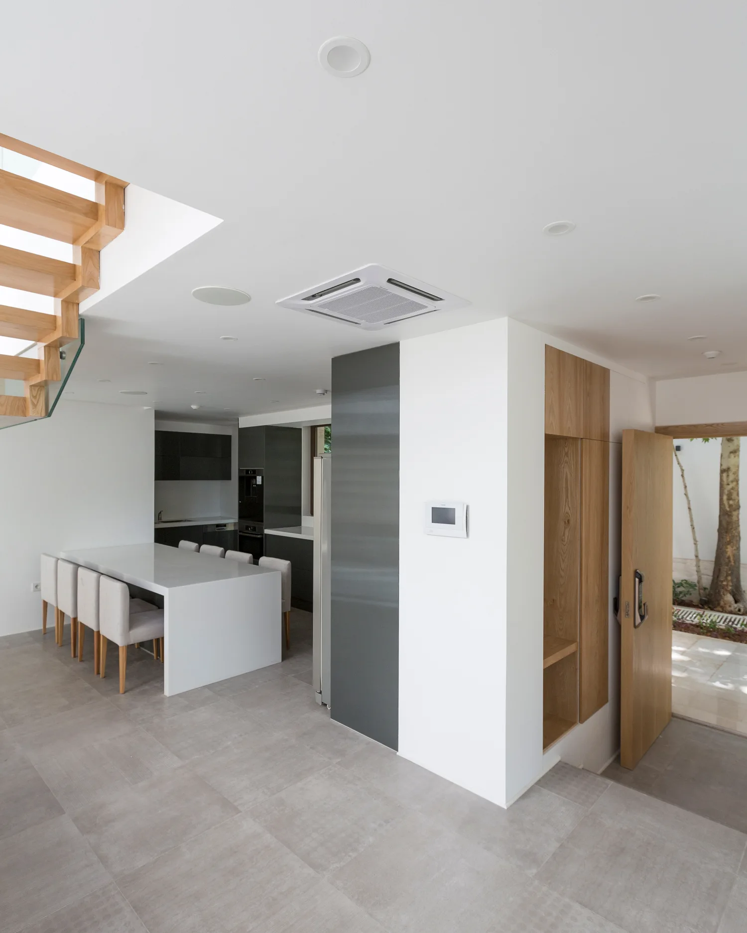 A minimalist open-concept kitchen features a large white dining island with light chairs, a wooden floating staircase, and gray tile floors.