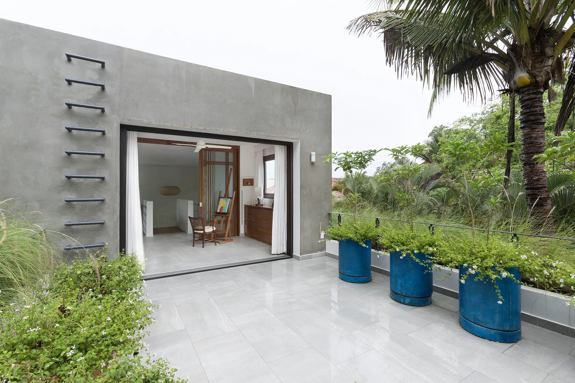 A modern concrete terrace features large-format grey floor tiles, three tall blue planters, and an open sliding glass door leading to an indoor studio.