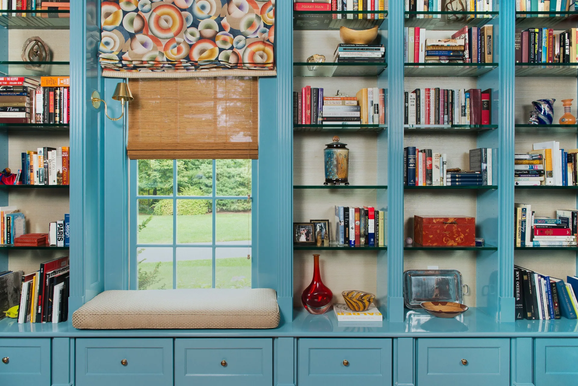 A vibrant blue built-in library bookcase surrounds a cushioned window seat, displaying colorful books on glass shelves.