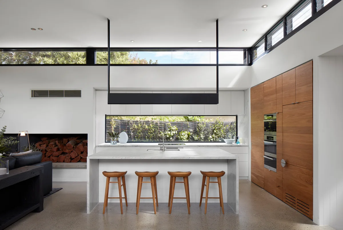 A modern kitchen with a white marble waterfall island, four wood stools, a long horizontal splashback window, and tall walnut cabinets.