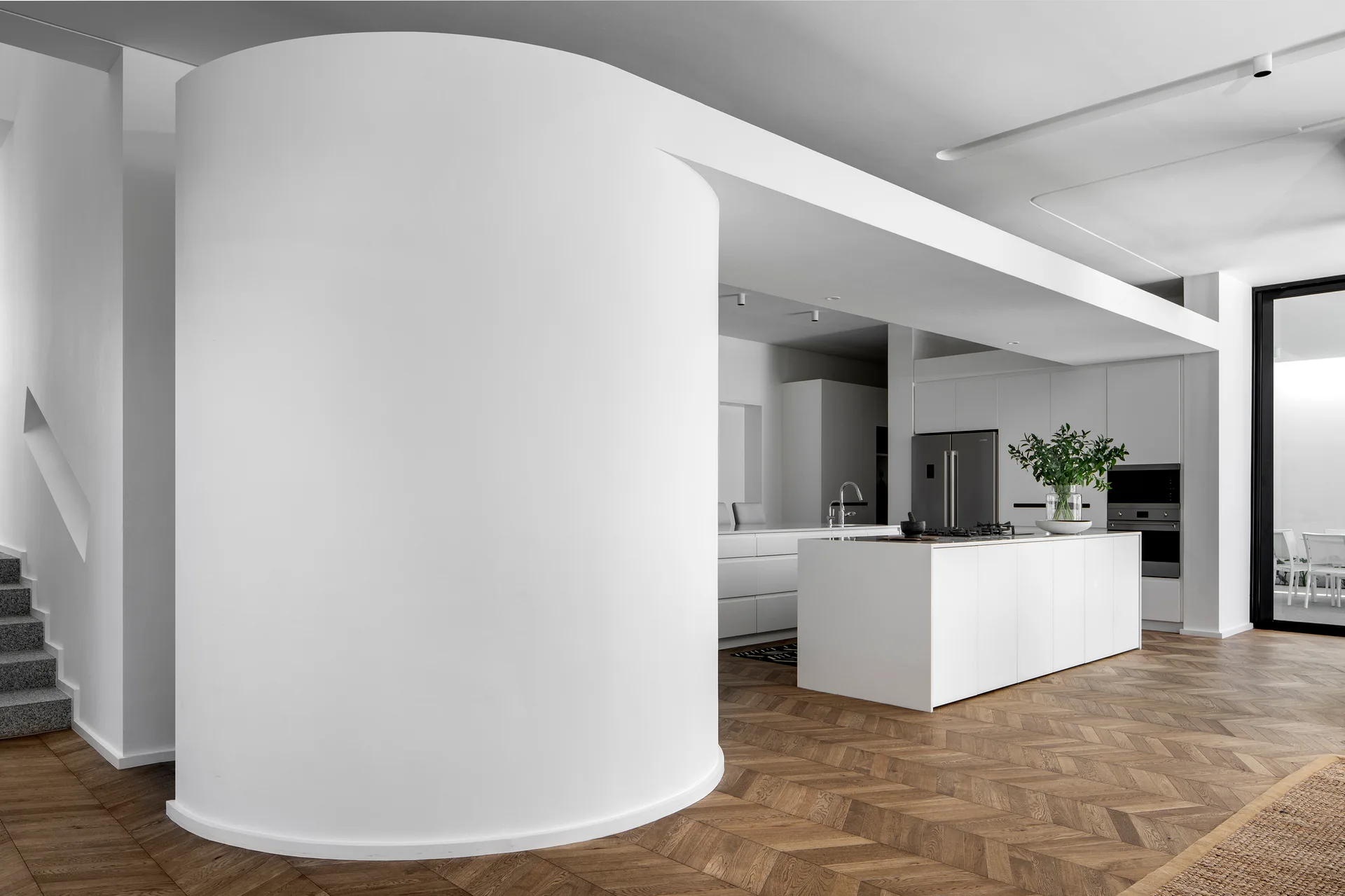 A minimalist kitchen features a large curved white partition wall, herringbone wood flooring, a central island with a cooktop, and flat-front cabinetry.