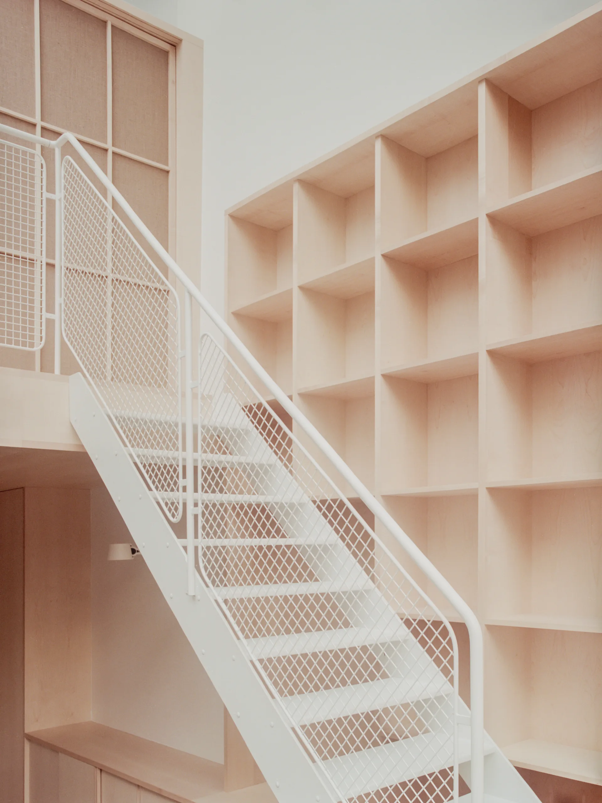 A light wood built-in grid shelving unit stands alongside a central white metal staircase with a mesh railing in a minimalist interior.