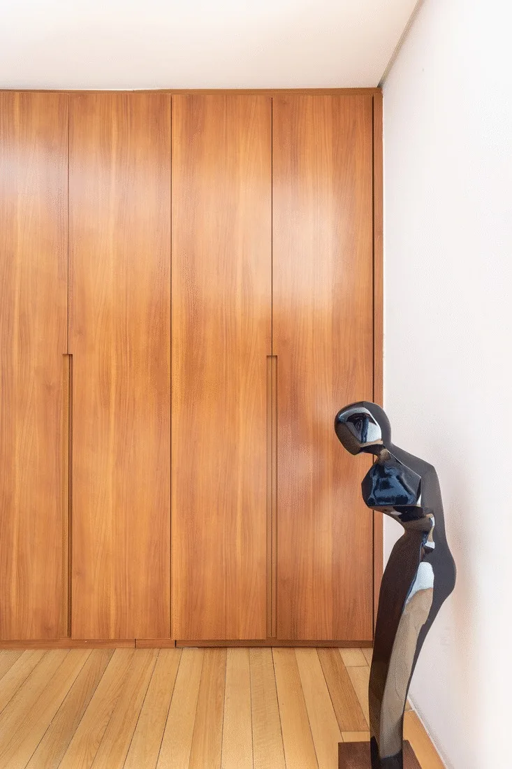 Floor-to-ceiling warm wood wardrobe cabinets with integrated vertical pulls stand next to a white wall and a black abstract sculpture on light wood floors.