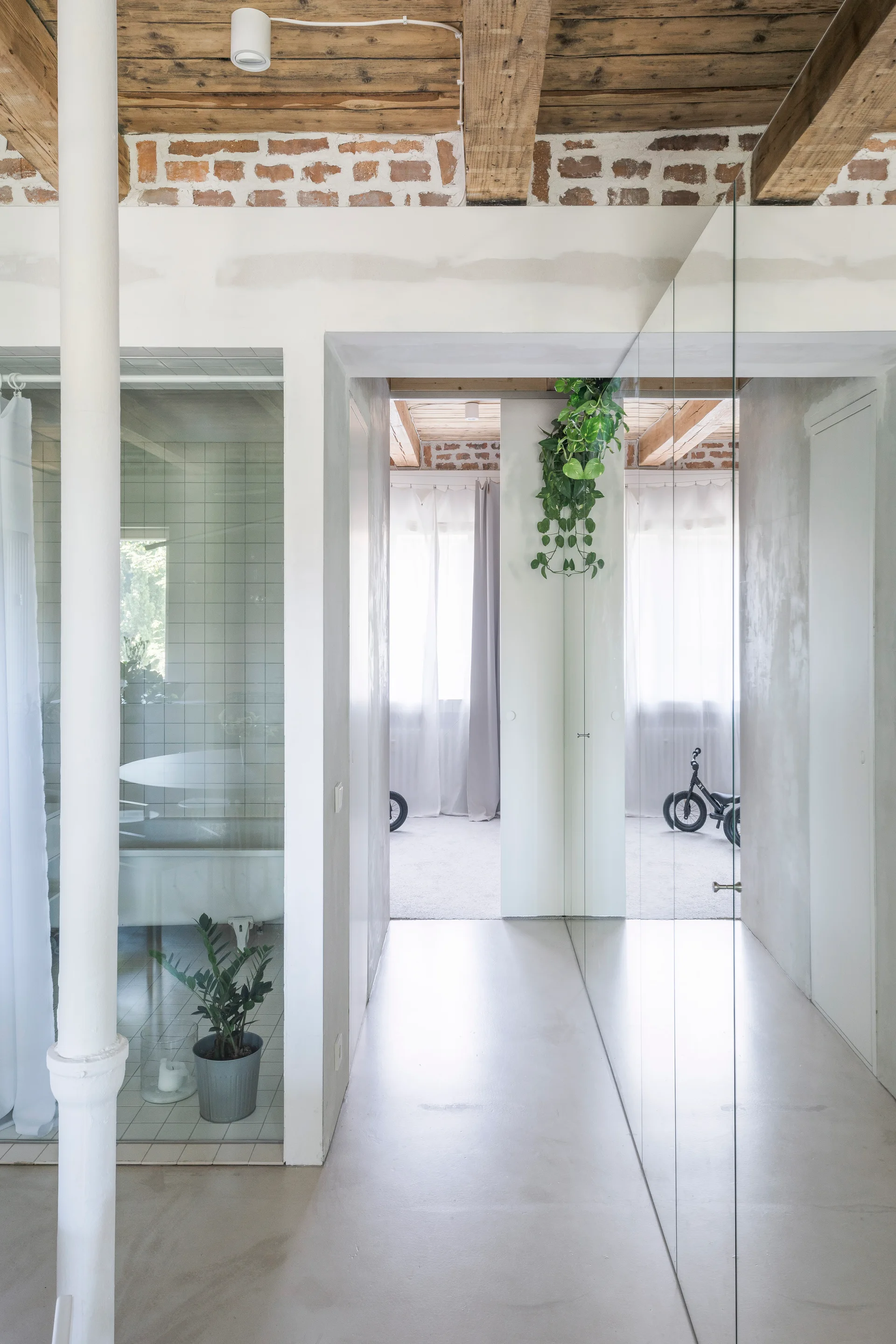 A white industrial hallway featuring exposed wood ceiling beams, a full-height mirrored wall, and a glass partition looking into a bathroom.