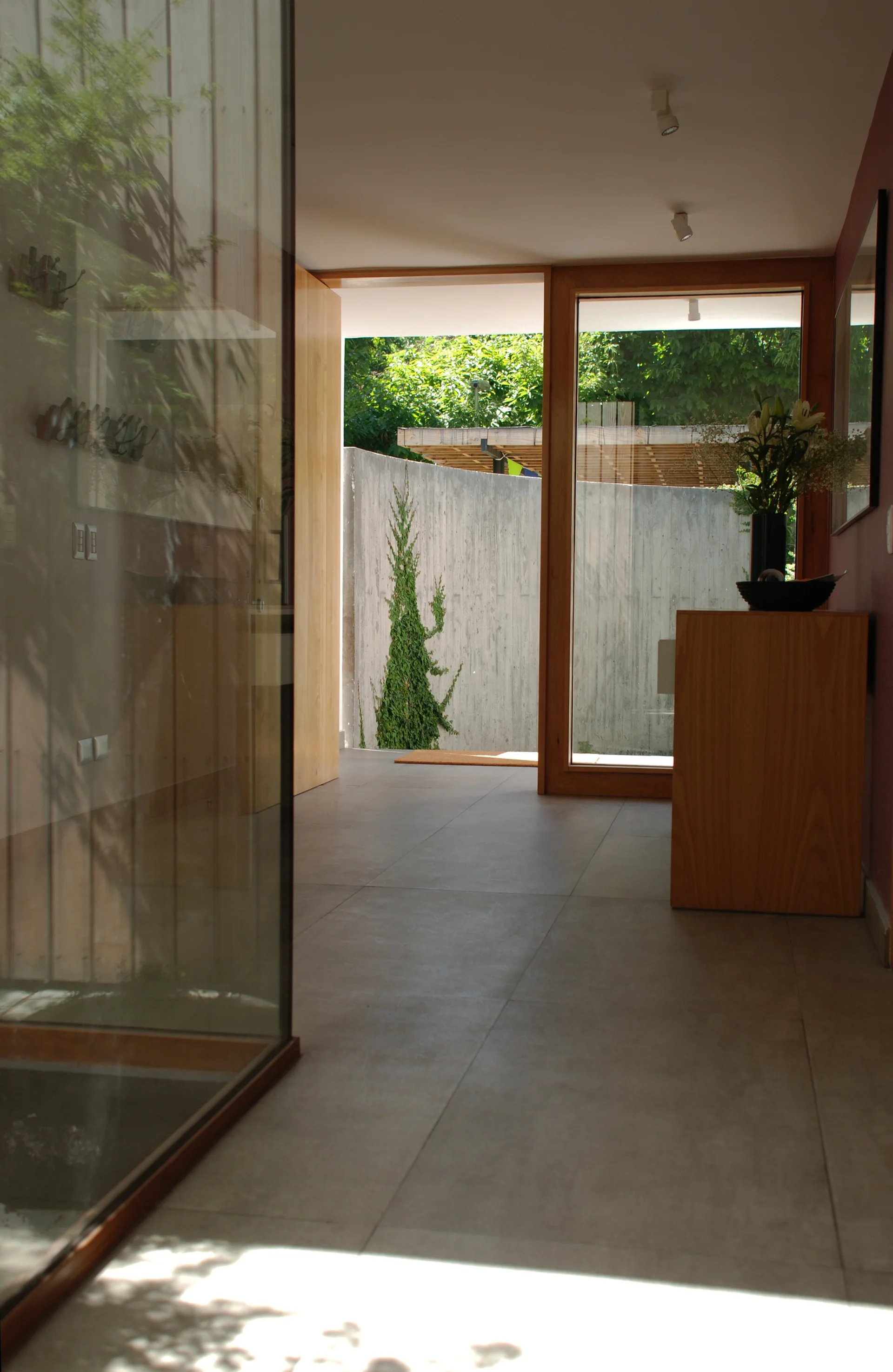 A modern minimalist entryway features large grey floor tiles, a wooden console cabinet, and a large glass door looking out to an exterior concrete wall.