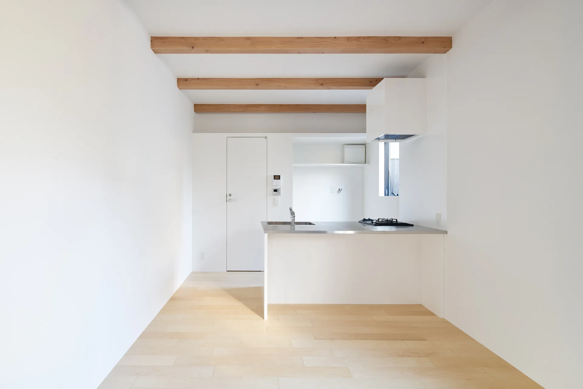 A minimalist white kitchen features a stainless steel countertop island, matte white walls, light wood flooring, and exposed wooden ceiling beams.