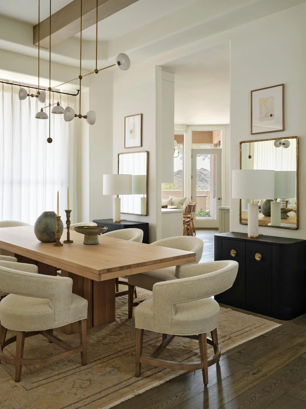 A bright dining room features a light wood table surrounded by curved cream chairs, flanked by two black sideboards with matching gold mirrors.