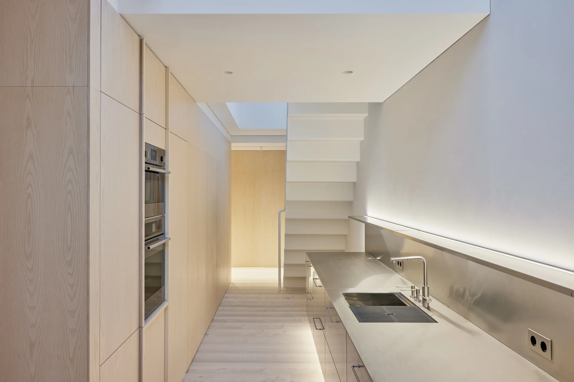 A minimalist kitchen features sleek light wood cabinets, a long stainless steel countertop, and a white staircase.