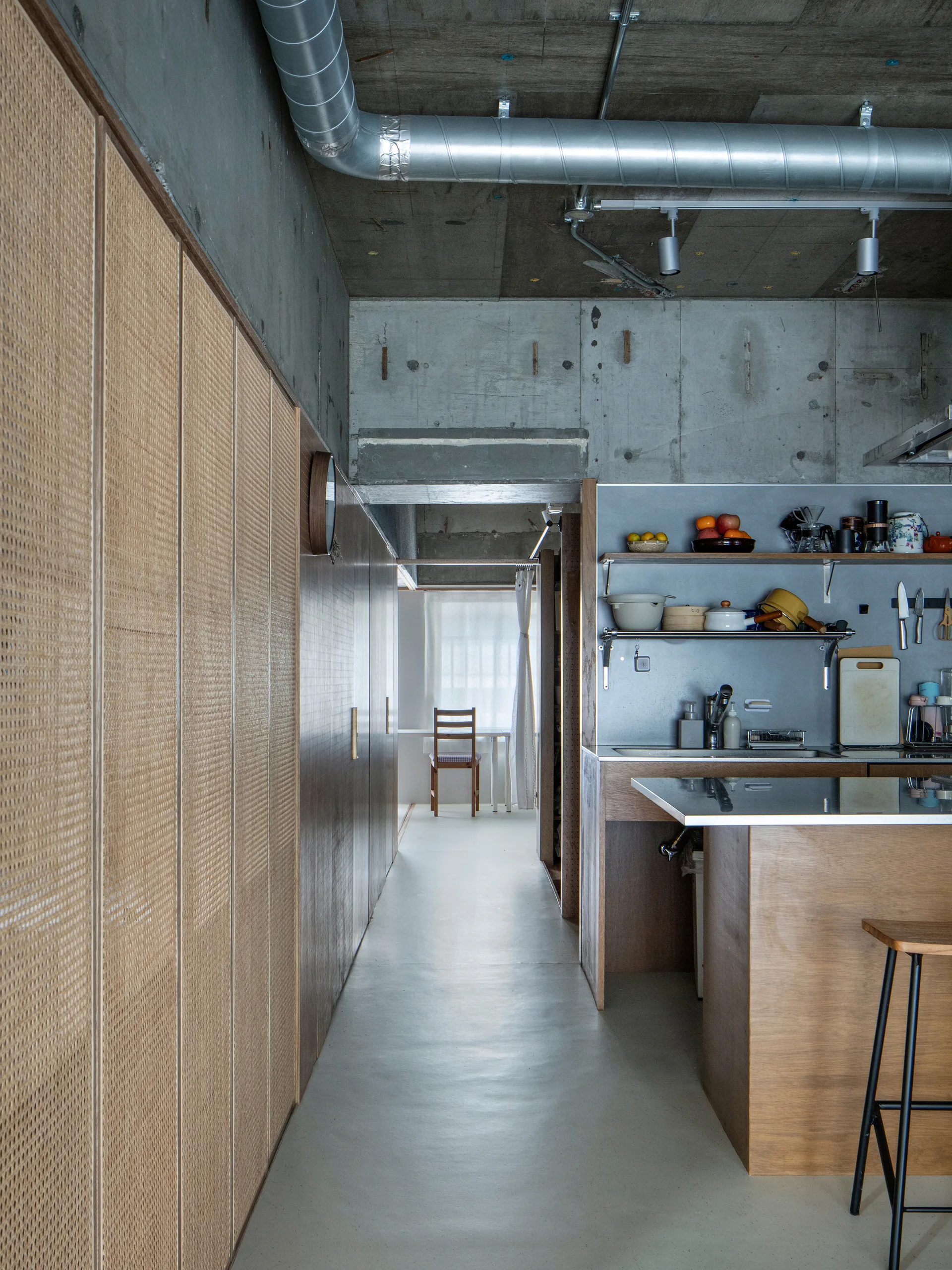 An industrial kitchen and hallway featuring exposed concrete walls and ceiling, a metal ventilation duct, and full-height rattan storage cabinets.