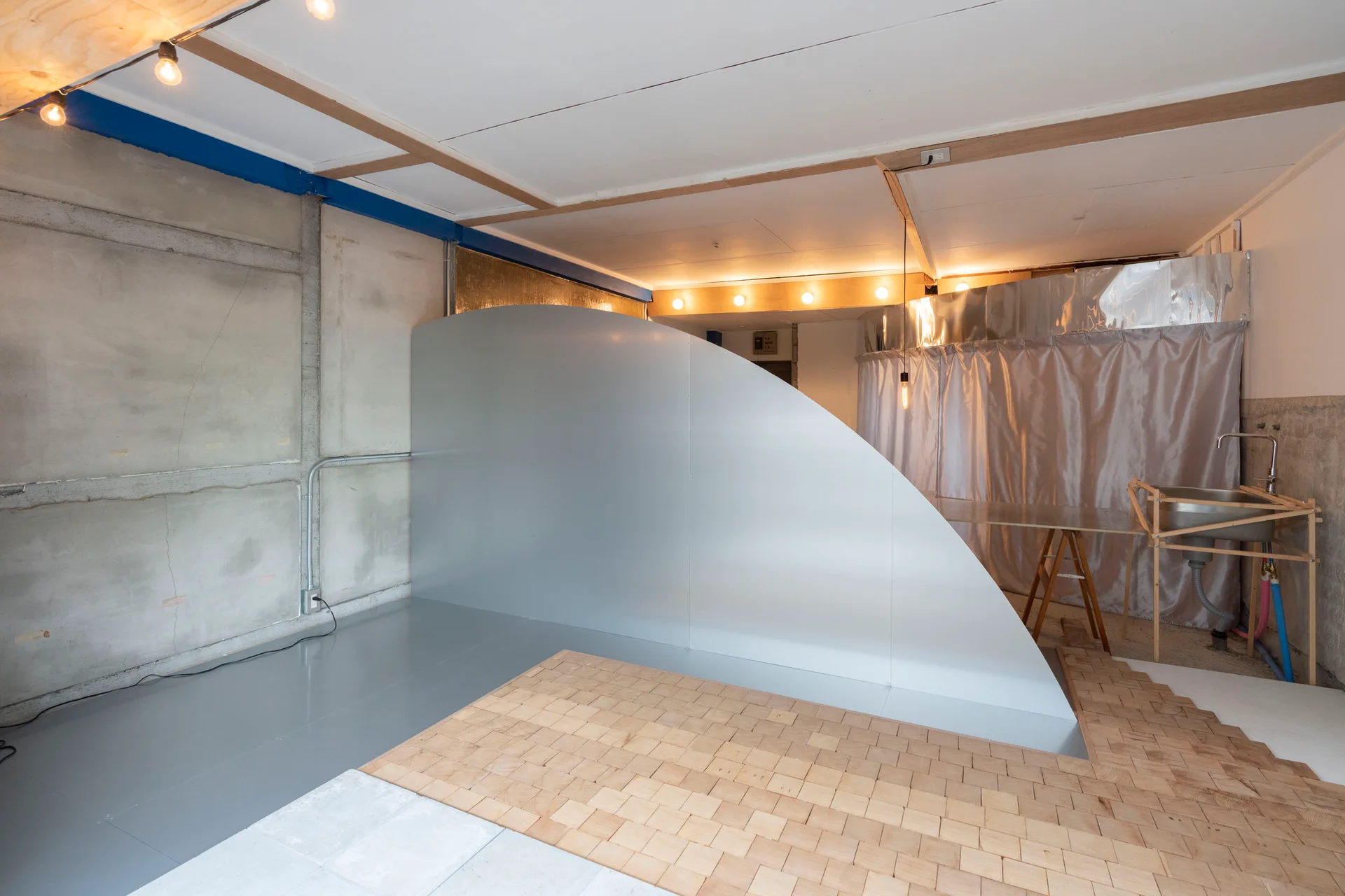 An industrial room featuring a large curved silver partition, raw concrete walls, exposed ceiling beams, and a light wood block floor section.