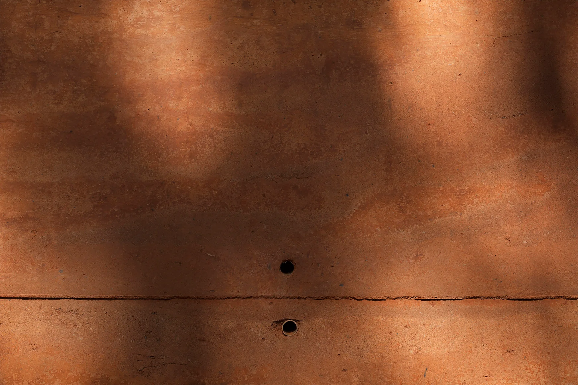 A textured terracotta rammed earth wall features a matte orange-red finish with horizontal layers, two circular holes, and dappled sunlight.