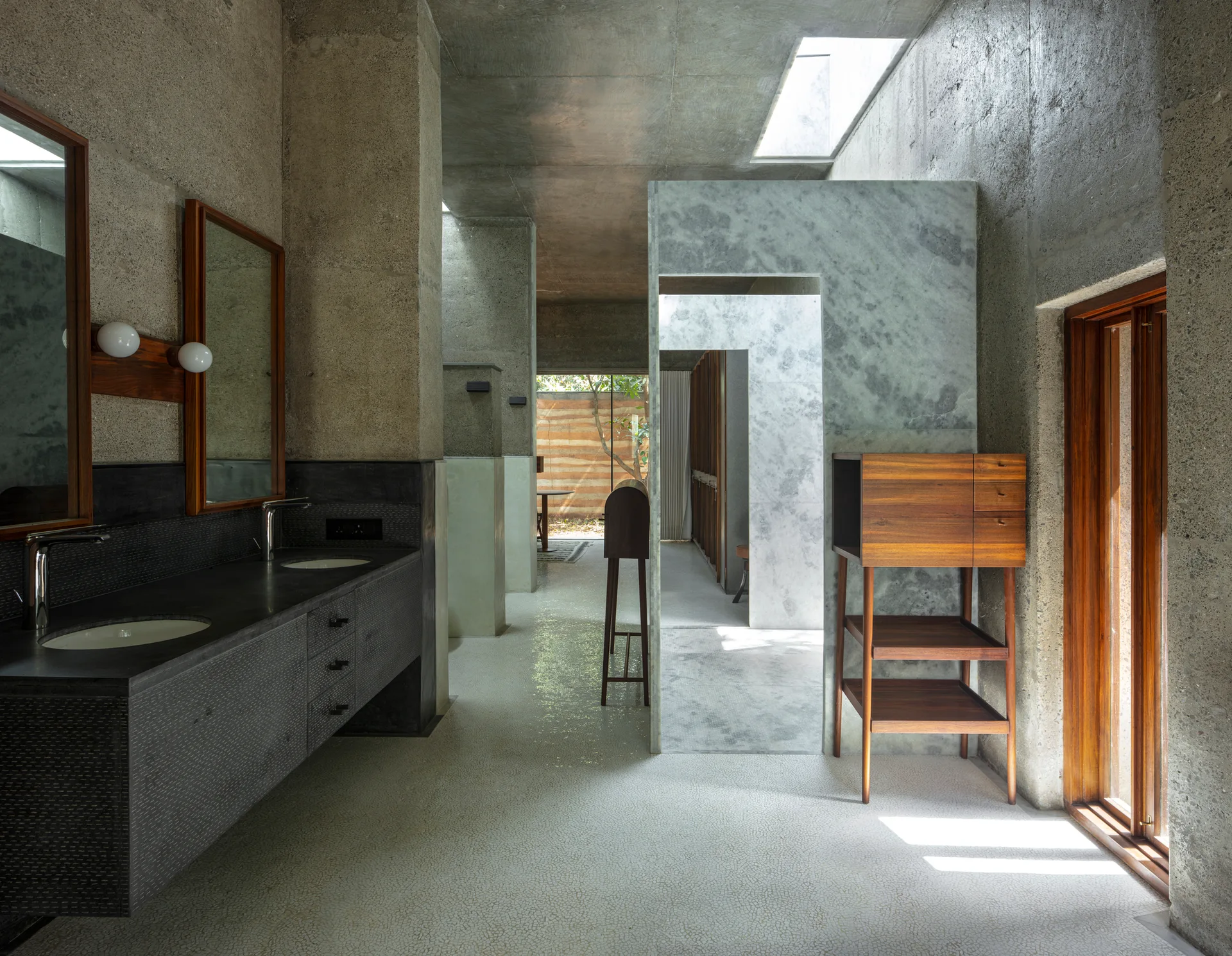 A spacious bathroom featuring raw concrete walls, a dark grey double vanity, stone panel partitions, and a warm wood cabinet near a window.