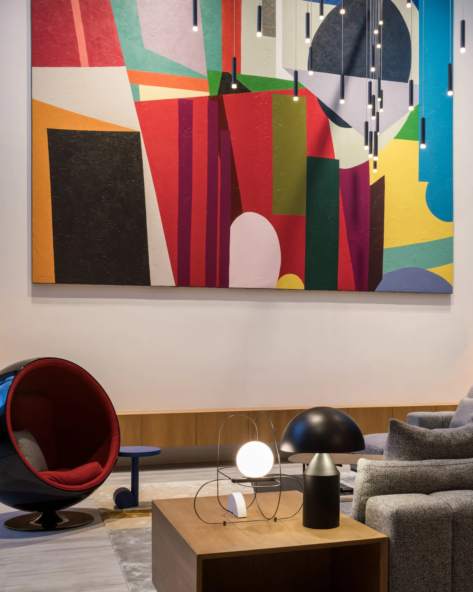 A modern living room displays a large colorful abstract painting above a floating wooden console, with a red ball chair and grey sofa nearby.