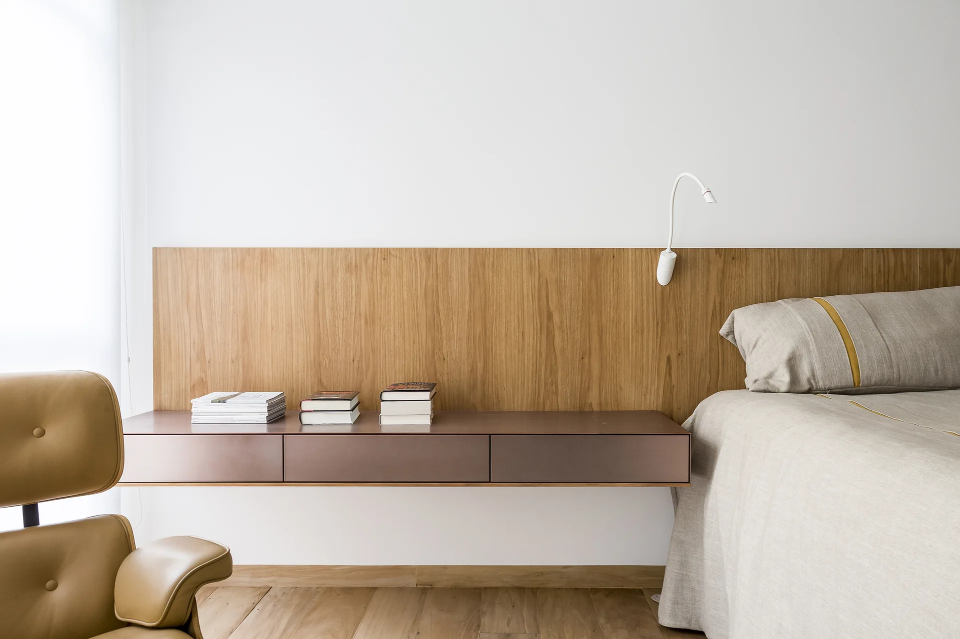 A minimalist bedroom featuring an extended wood panel headboard, a wall-mounted floating storage drawer with books, and a tan leather chair.