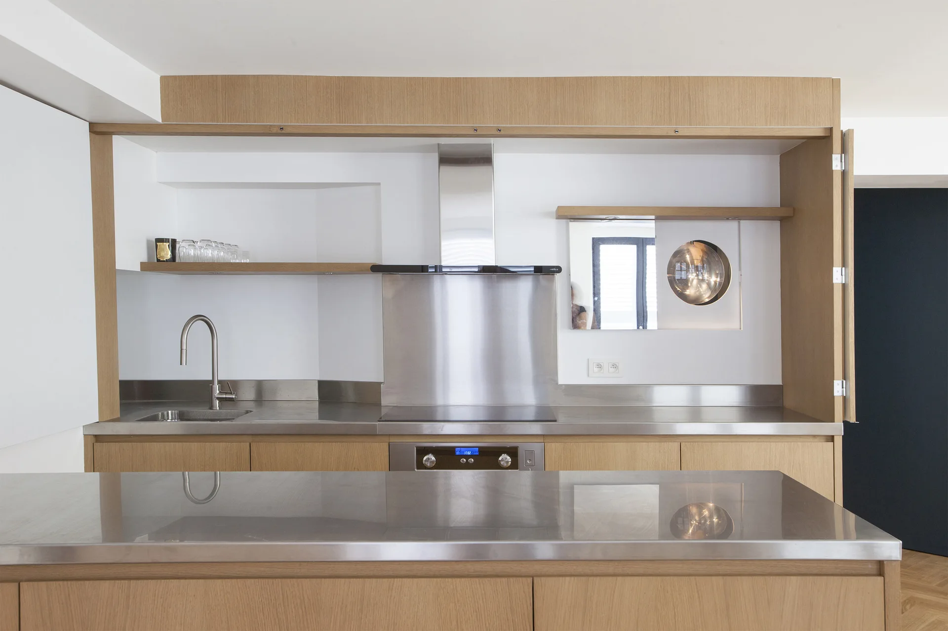 A minimalist kitchen with light wood cabinets, a stainless steel countertop and backsplash, an integrated cooktop, and floating shelves.