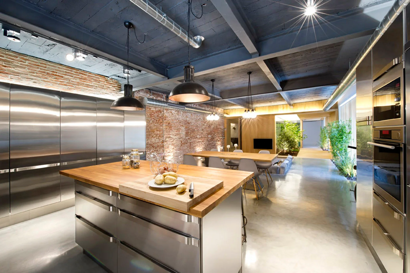 An industrial kitchen features a stainless steel island with a wooden countertop, built-in metal cabinets, concrete flooring, and an exposed brick wall.
