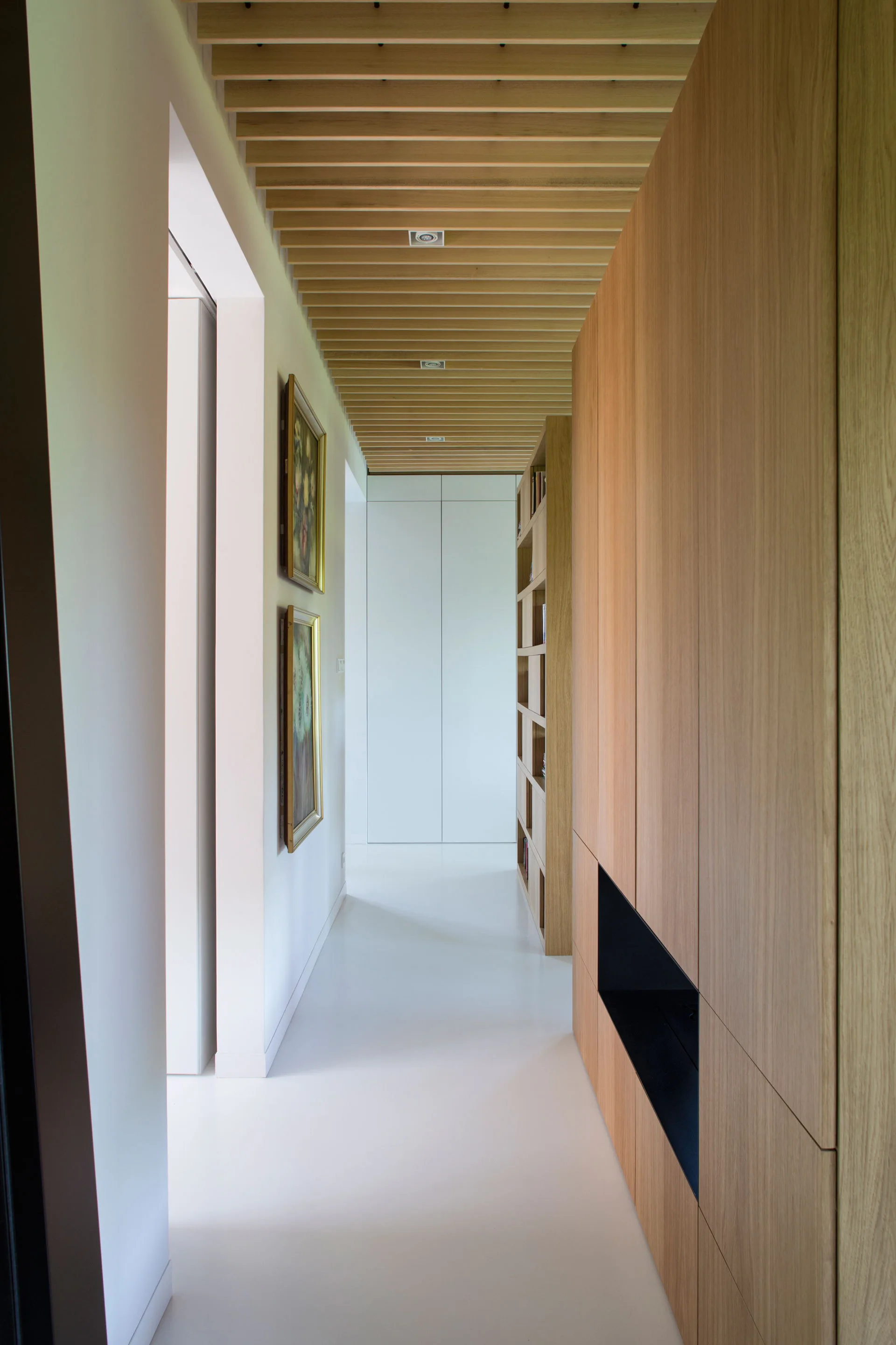A minimalist white hallway featuring a light wood-slatted ceiling, custom full-height wooden cabinetry, and a clean white floor.