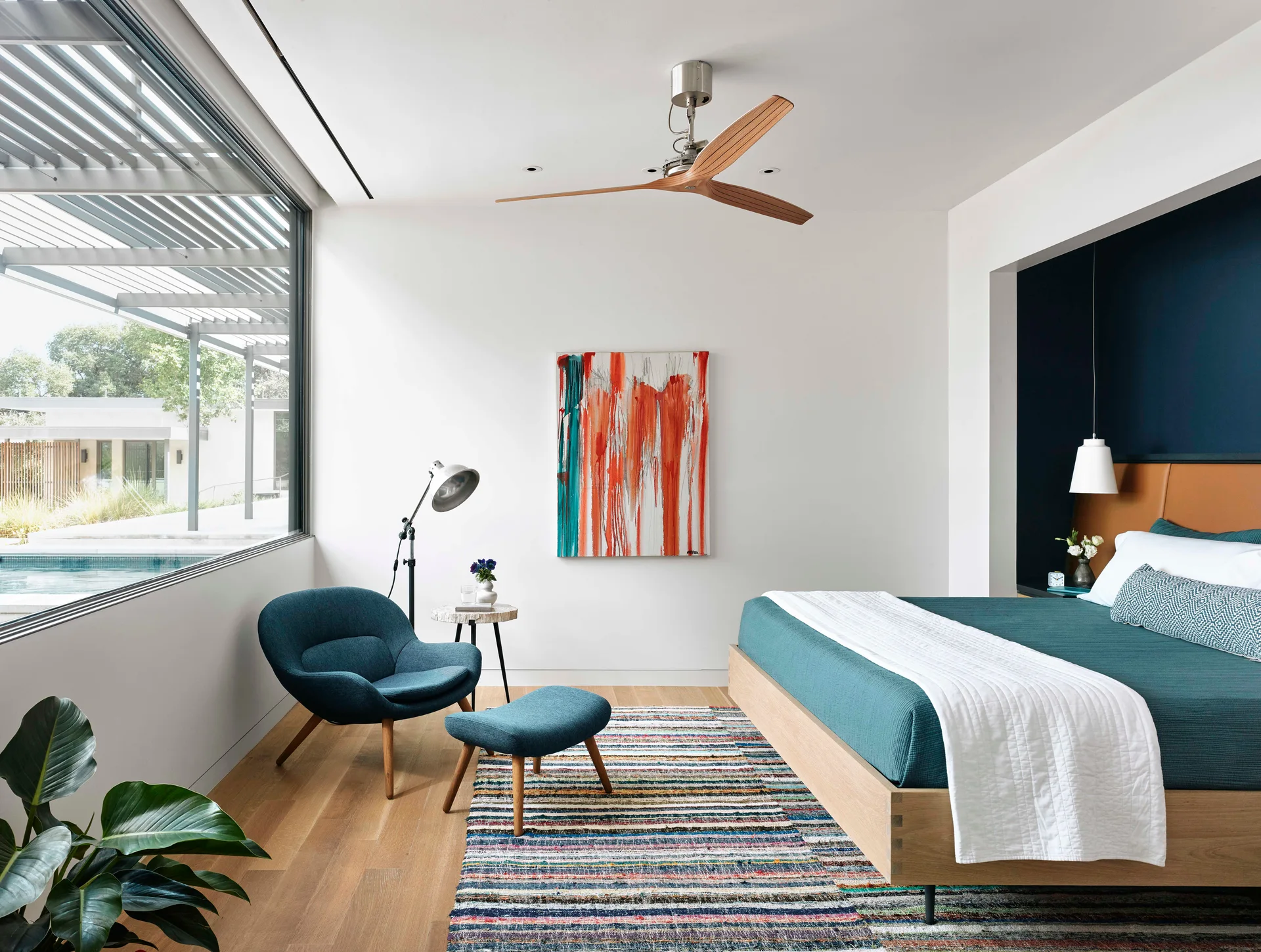 A modern bedroom features a teal bed set within a dark navy alcove, a matching teal armchair, and a colorful striped rug on wood floors.