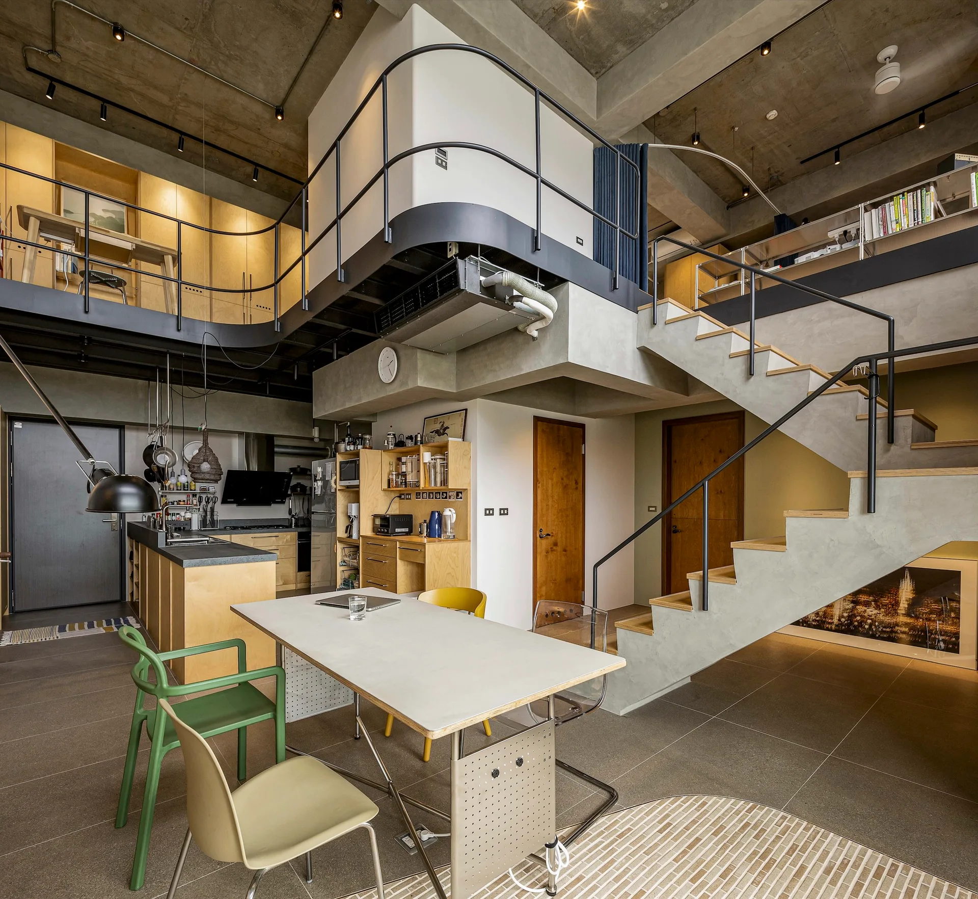 An industrial loft interior with a concrete staircase leading to a mezzanine, next to a dining area with mismatched chairs and a wood kitchen.