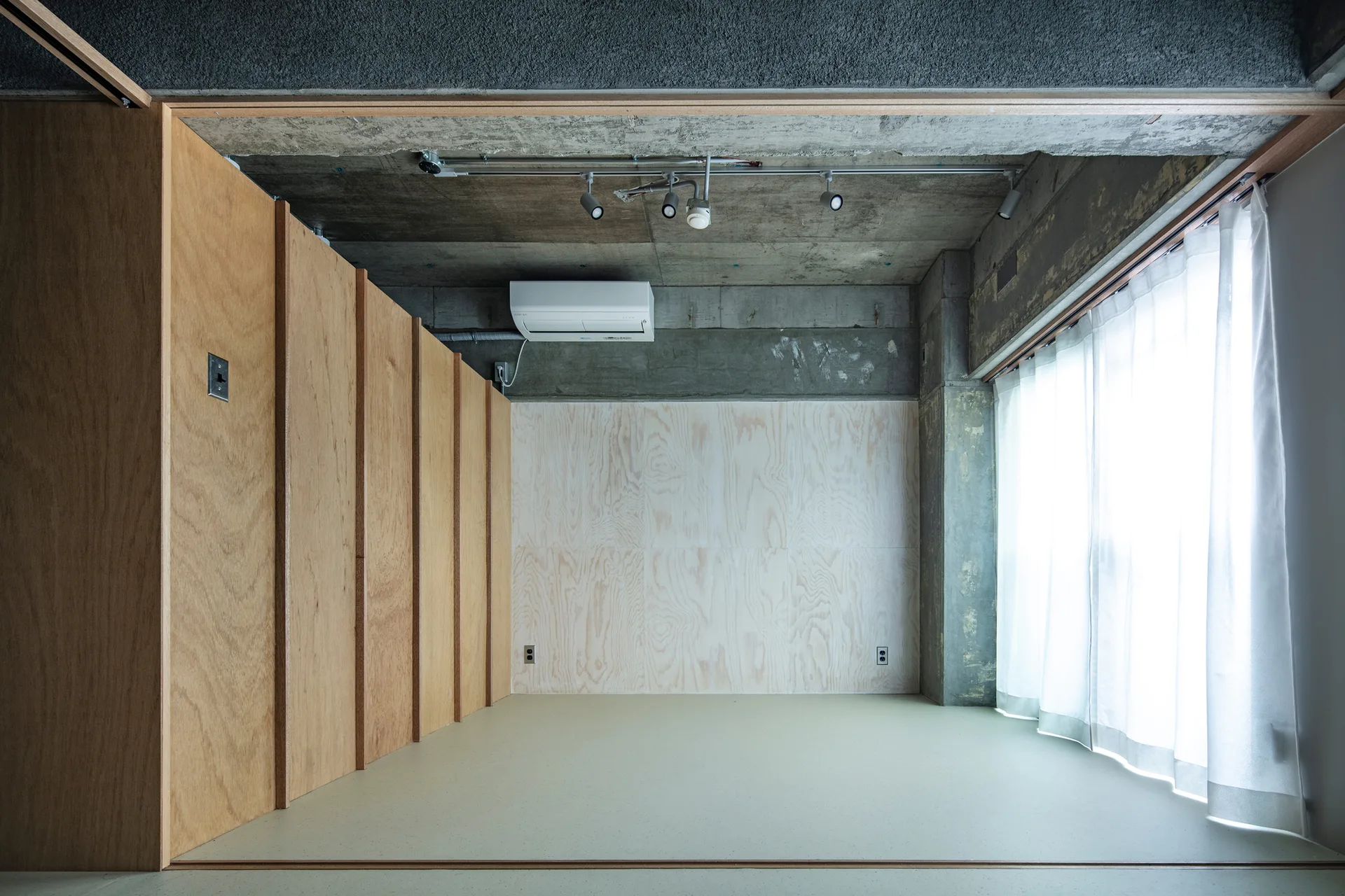 A minimalist room features a raw concrete ceiling, light plywood paneled walls, and white curtains framing a window.