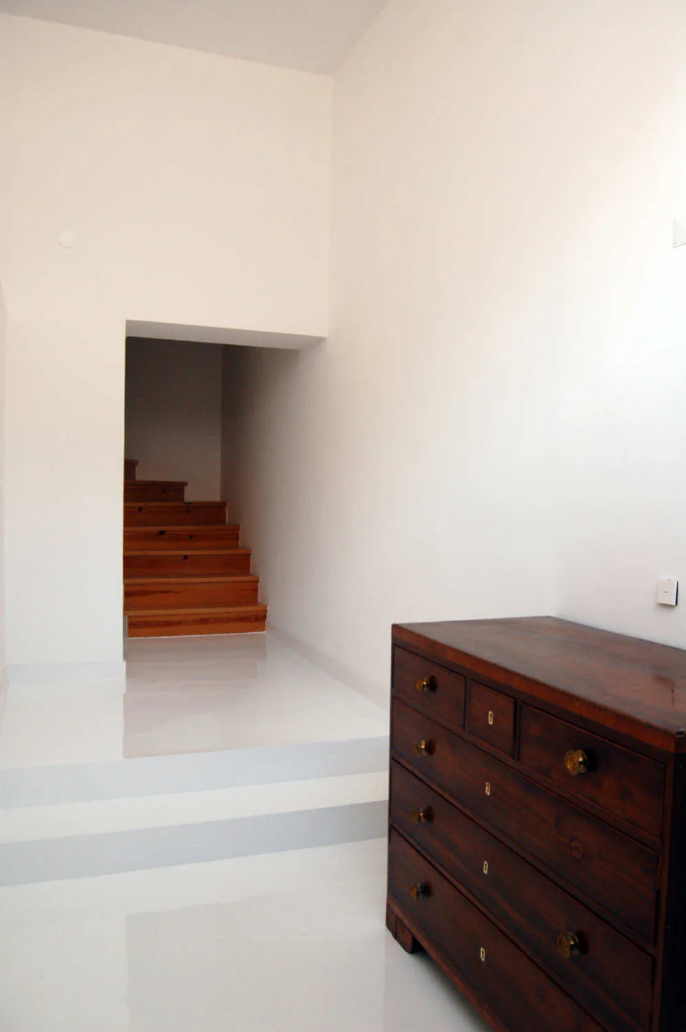 A dark wood chest of drawers stands on a glossy white reflective floor in a minimalist white hallway leading to a wooden staircase.
