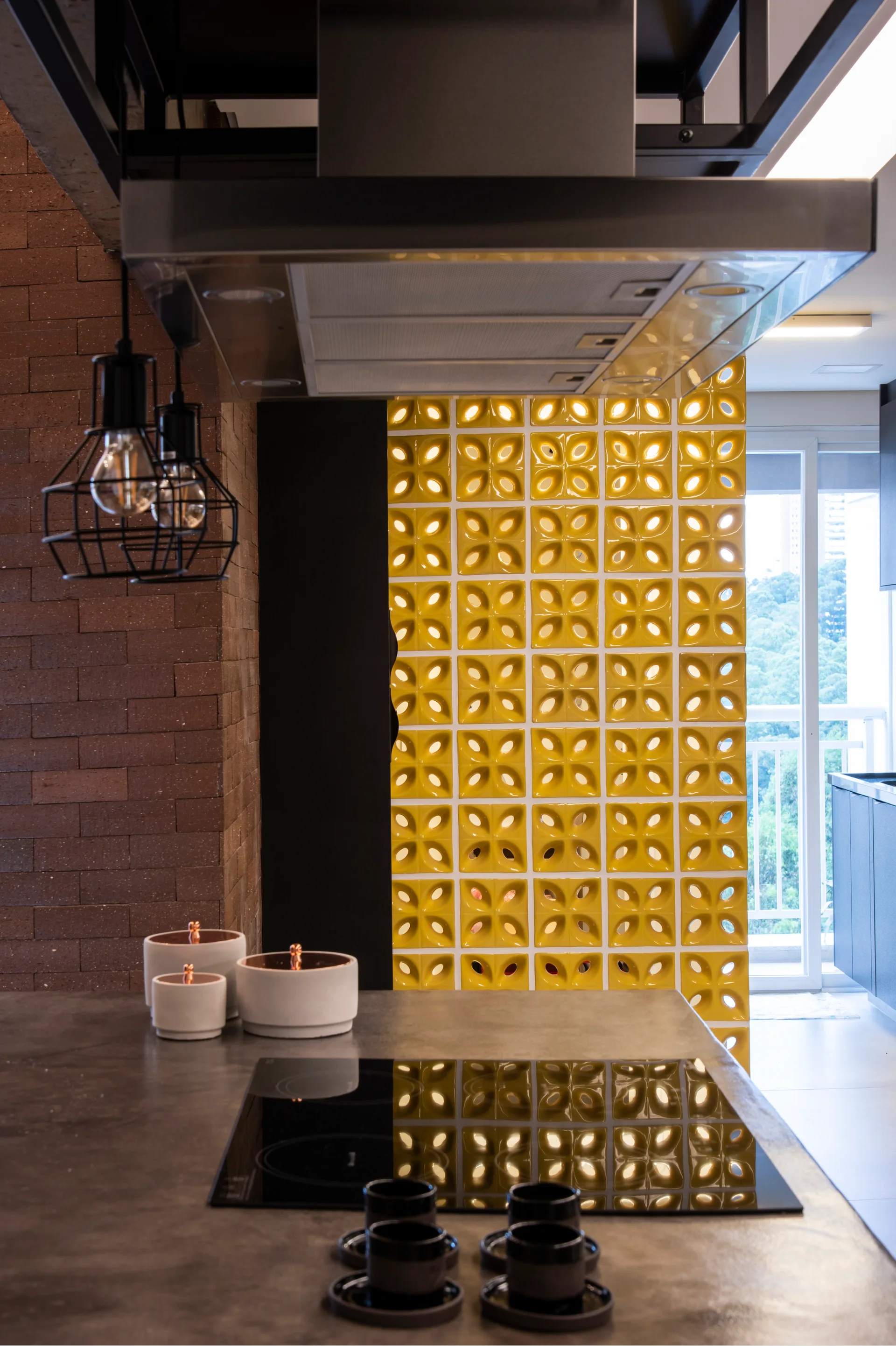 A kitchen island with an induction cooktop stands in front of a textured brick wall and a bright yellow patterned breeze block screen.