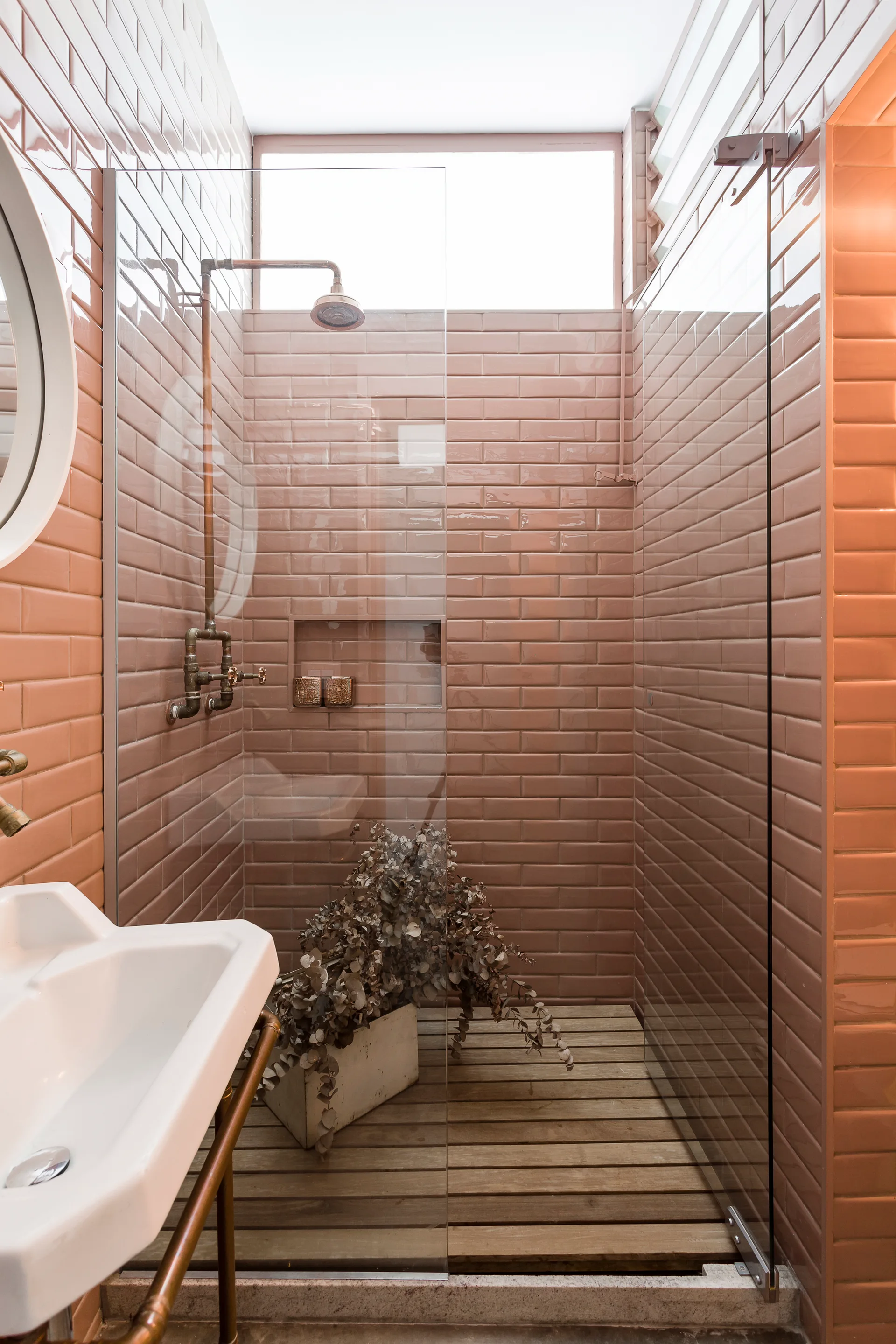 A modern bathroom features pink subway tiled walls, a glass shower partition, exposed copper plumbing, and a slatted wood shower floor.