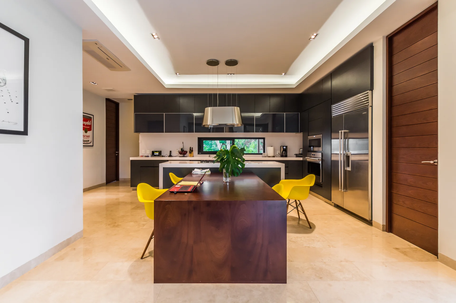 A modern kitchen with matte black cabinets, a dark wood island table with yellow chairs, and polished beige floor tiles under a recessed ceiling.