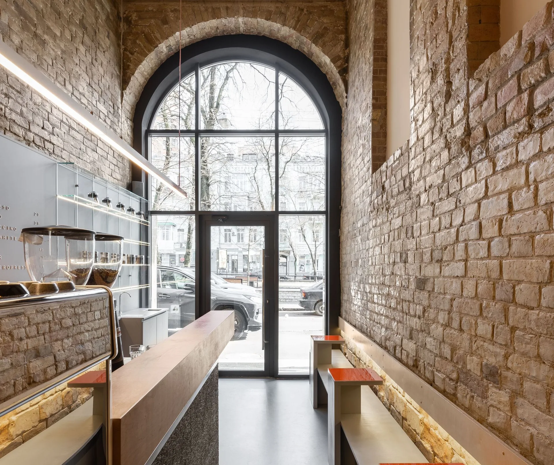 Interior of a narrow cafe with exposed brick walls, a wooden service counter, minimalist bench seating, and a tall arched window looking out to the street.