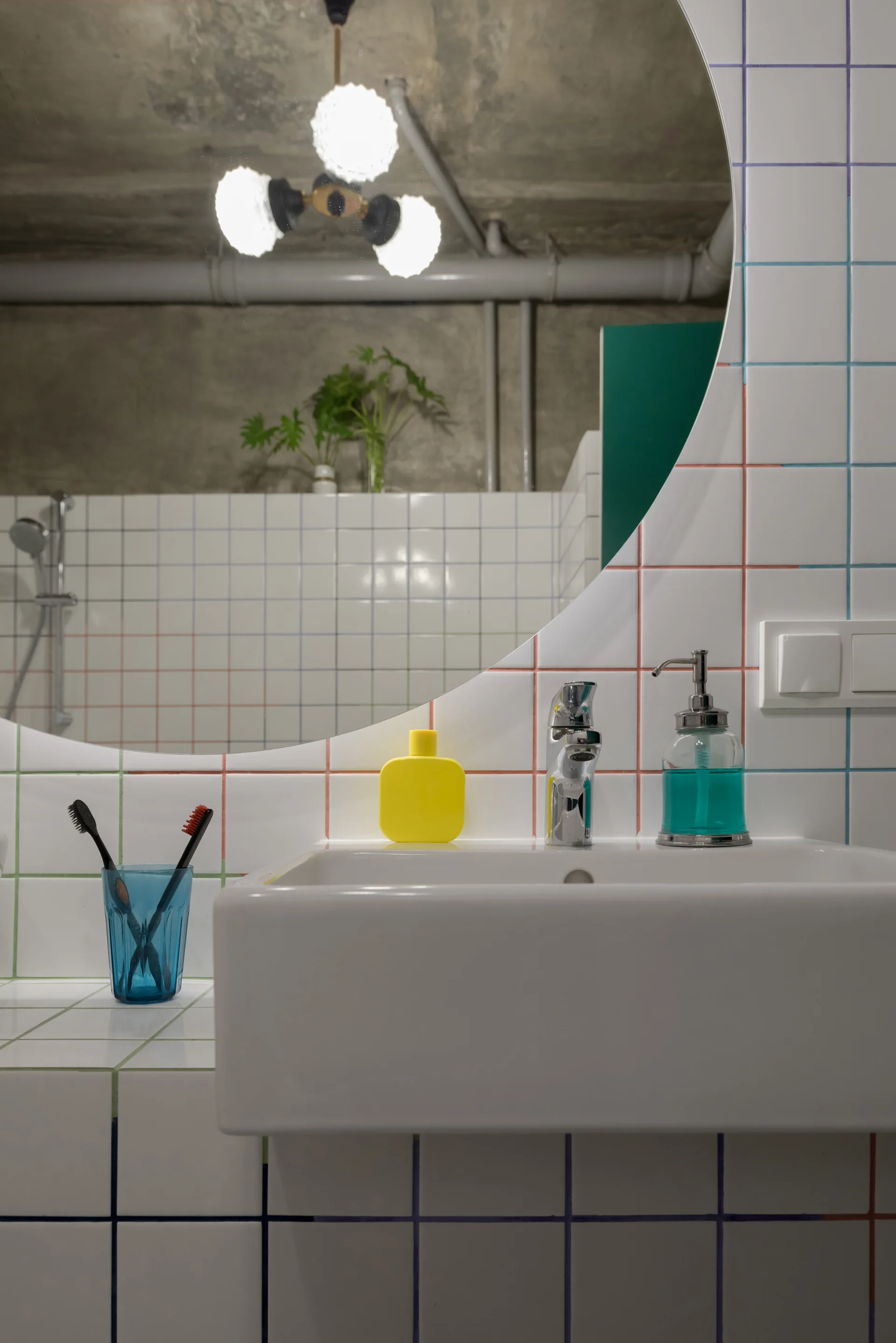 A white ceramic bathroom sink sits in front of a grid of square tiles featuring pink, blue, and green grout beneath a large round mirror.