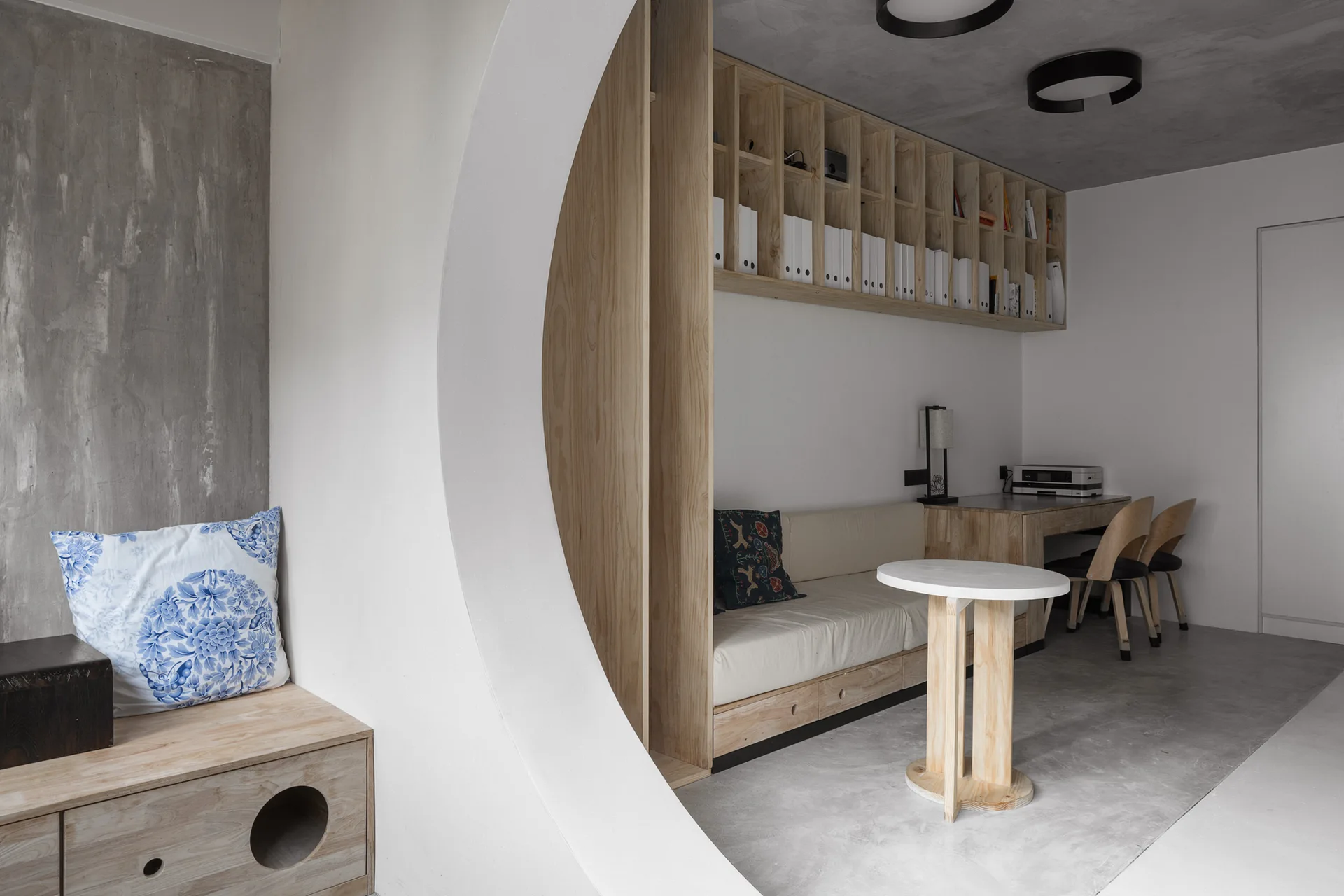 A modern home office with a large white circular partition separating a wooden desk and daybed from a grey concrete floor and ceiling.