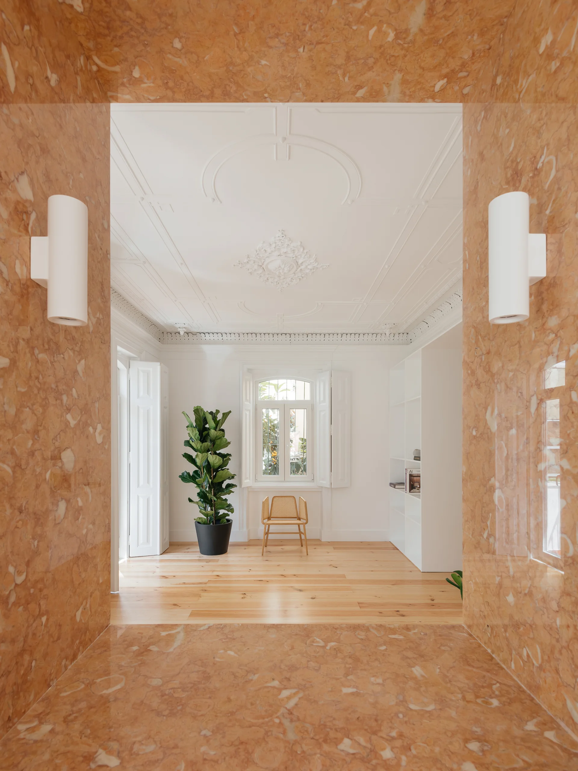 A bright minimalist room with a wood floor and ornate ceiling, viewed through a polished peach marble archway with white cylinder sconces.