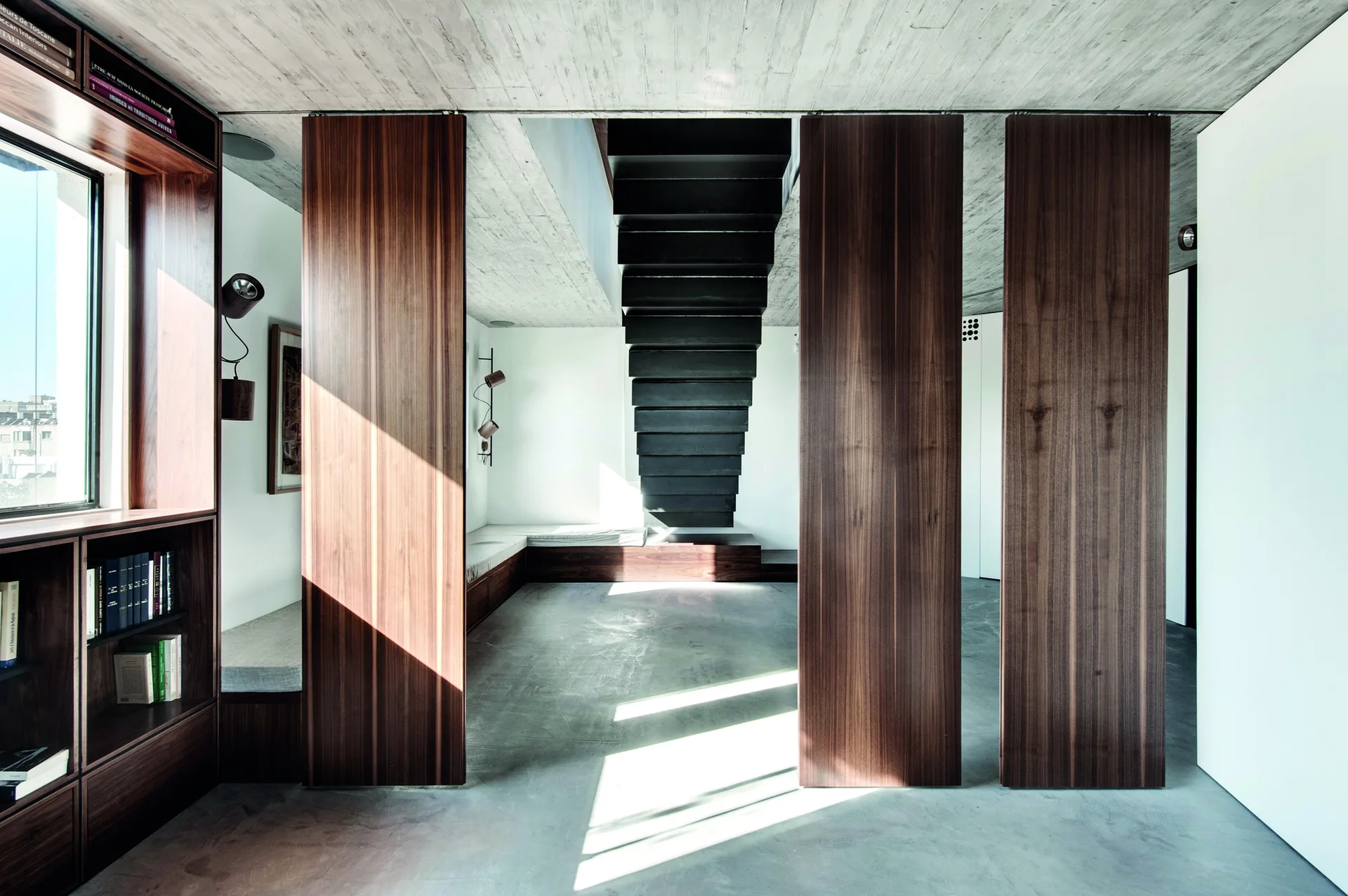Minimalist interior featuring a floating black staircase behind three large vertical wood panel dividers, concrete ceiling, and a built-in bench.