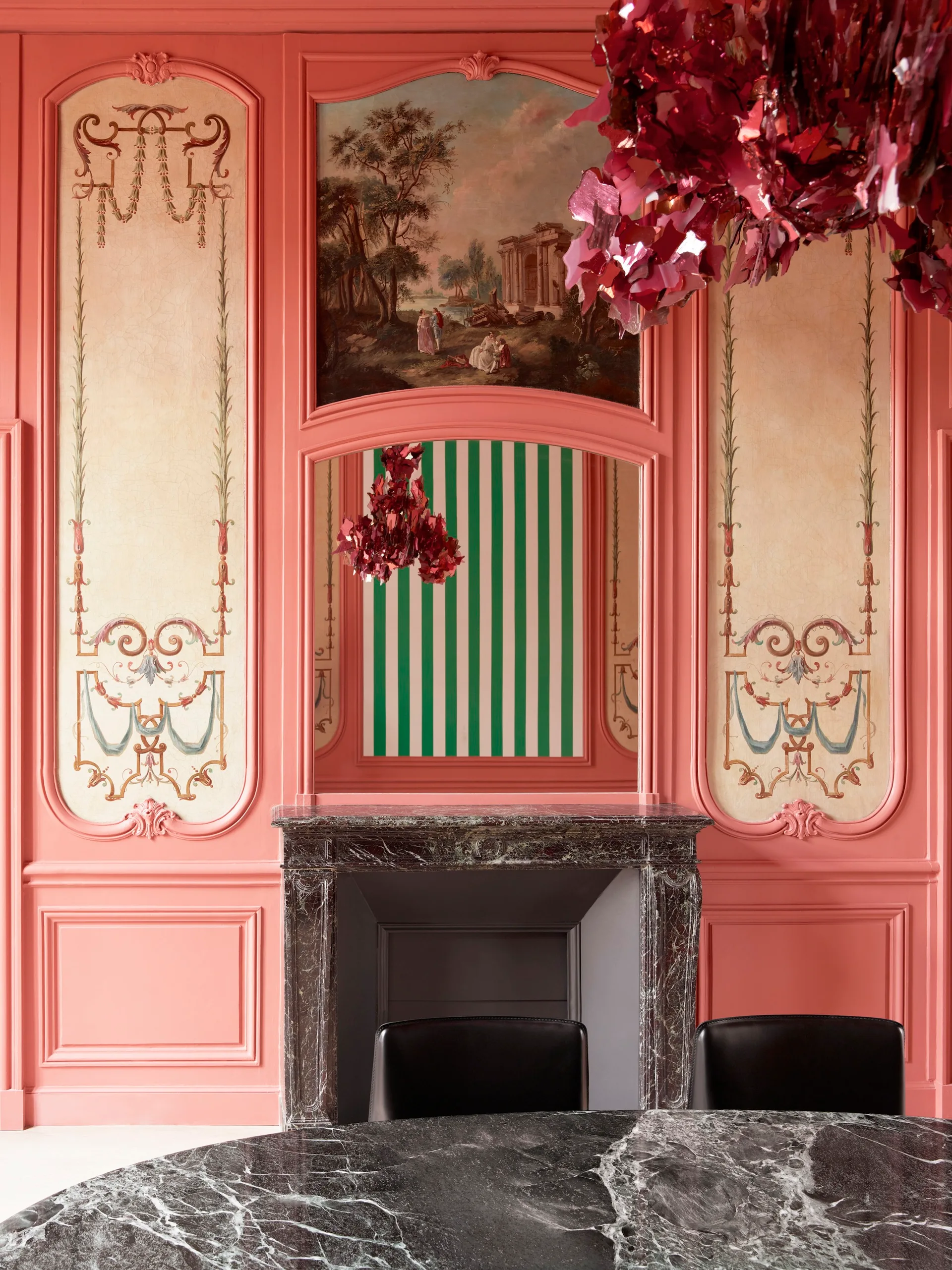Pink ornate wall panels surround a dark marble fireplace, featuring a painted pastoral mural and a mirror reflecting green and white stripes.