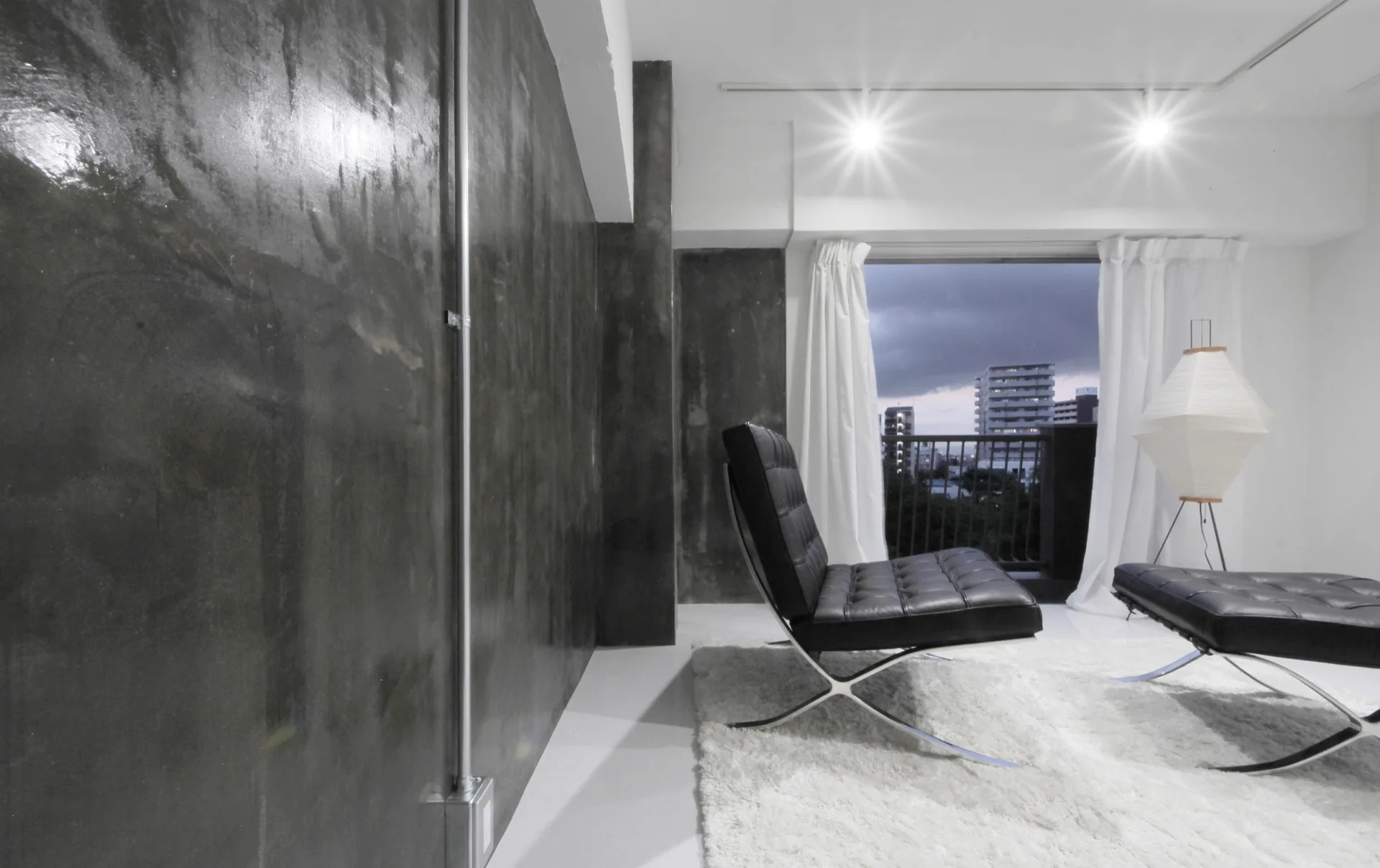 A black tufted leather lounge chair rests on a white shaggy rug next to a dark polished concrete wall with exposed conduit.
