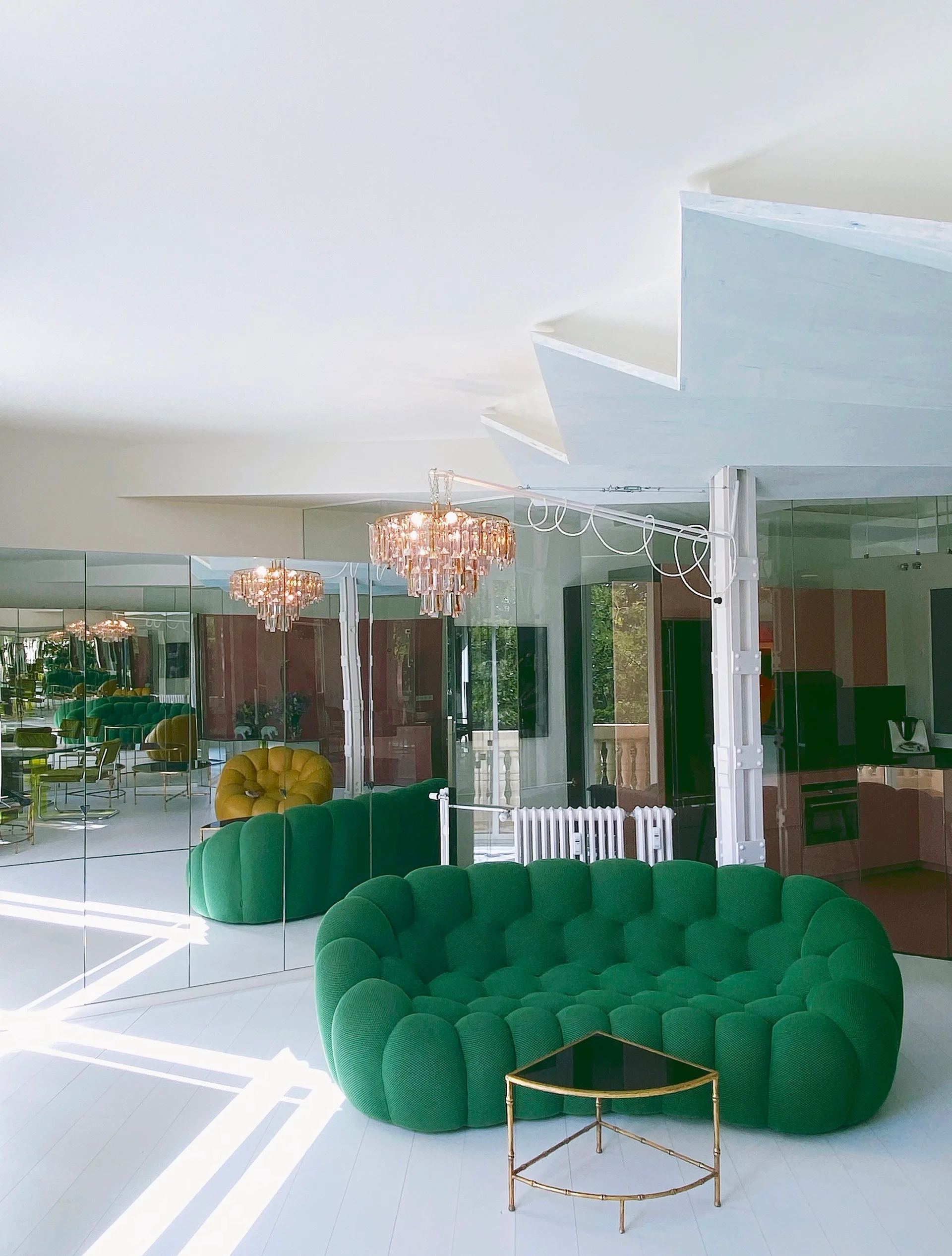 A bright living room featuring a green tufted sofa, a gold accent table, a tiered crystal chandelier, and walls covered in mirror panels.