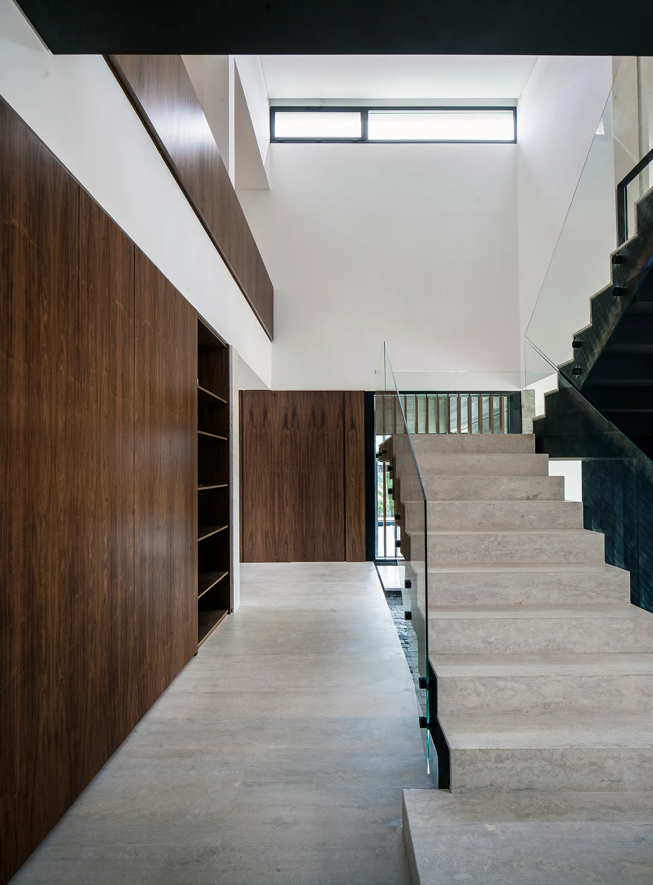 A minimalist double-height entryway features stone stairs with a glass railing opposite a full-height wood-toned storage wall.
